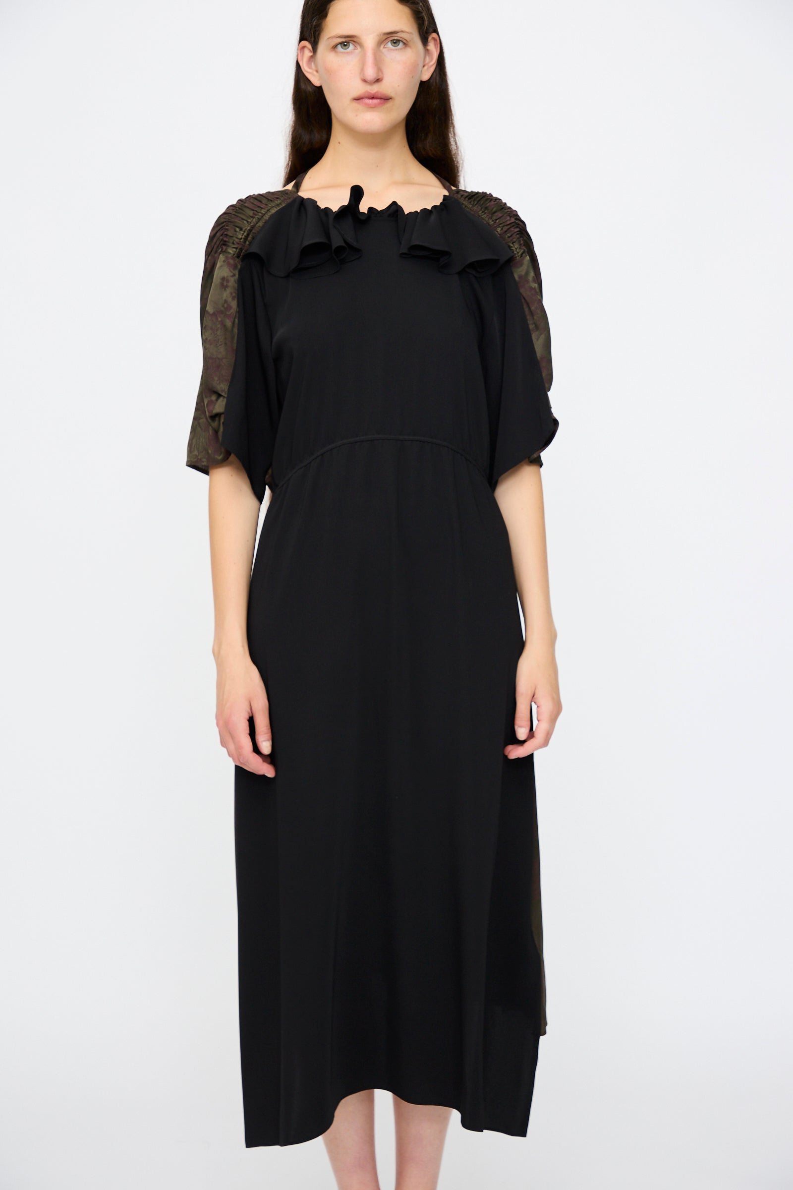 A woman faces forward, wearing the MM6 Midi Dress in Black and Khaki Burgundy. The dress features a floral print, short sleeves with textured details, a ruffled neckline, and a relaxed fit. The background is plain white.