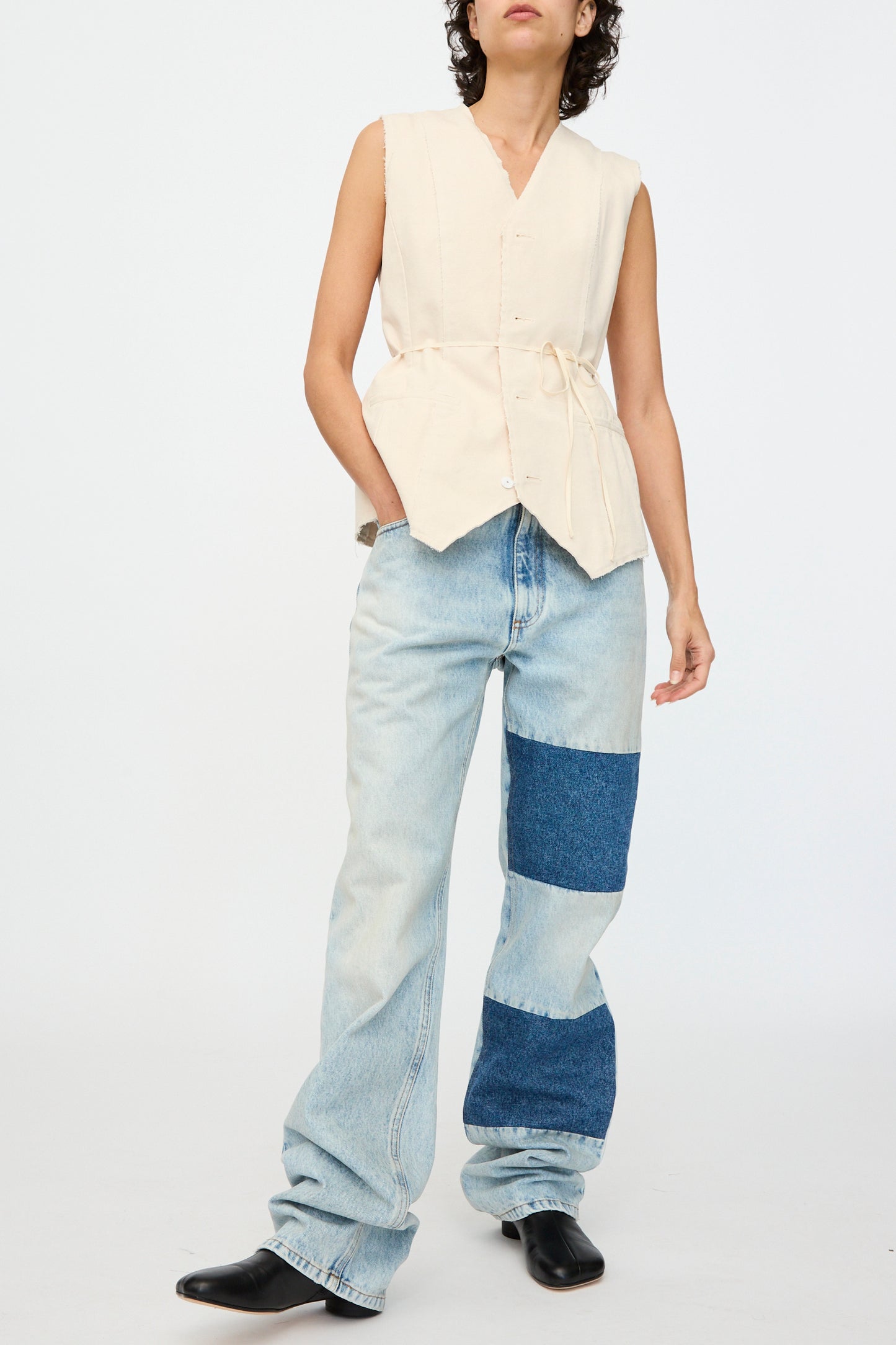 A person wears a sleeveless beige top with tied detail, paired with MM6's Pants 5 Pockets: relaxed fit, light blue jeans featuring dark blue patchwork denim. They complete the look with black shoes and stand against a plain white background.