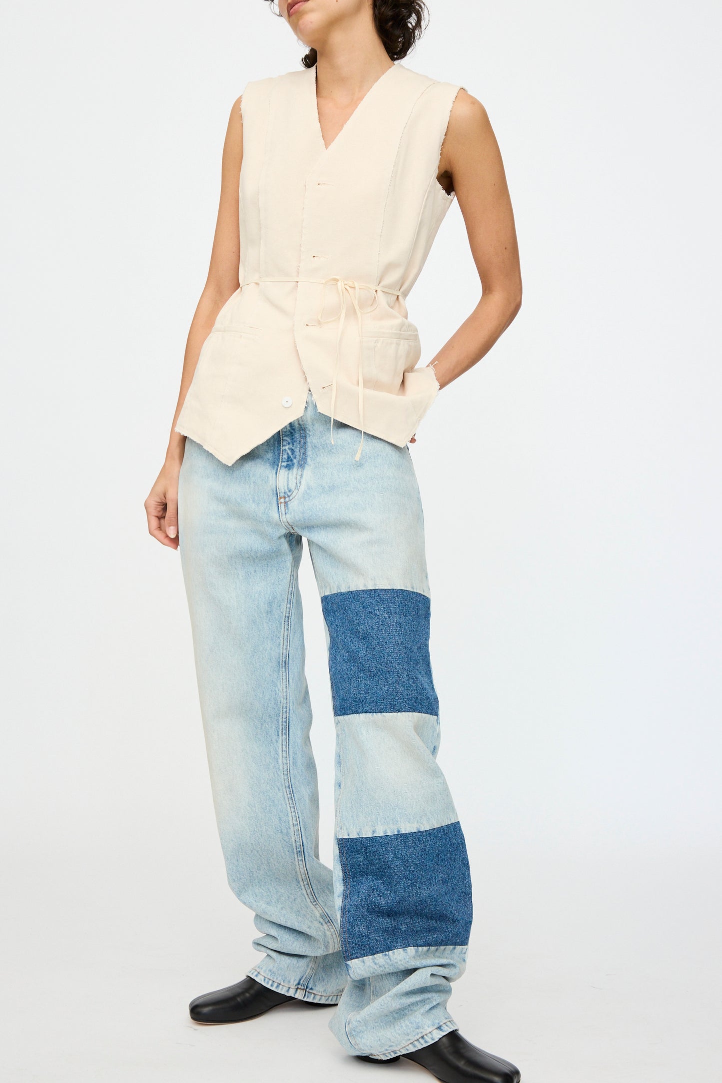 A person wearing a sleeveless beige top with drawstrings and MM6's Pants 5 Pockets, which are loose light blue jeans with a patchwork denim design, paired with black shoes.