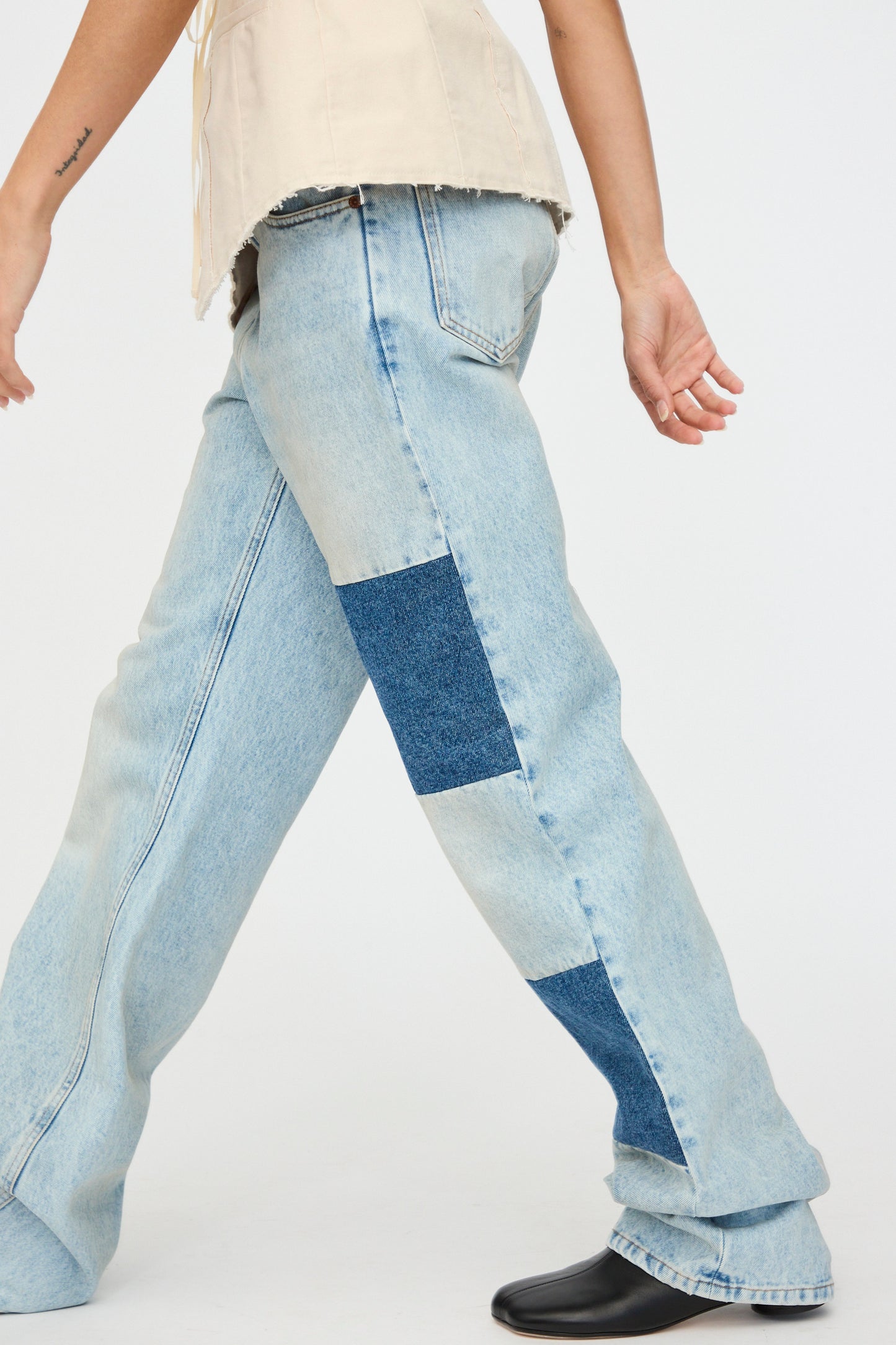 A person in MM6's relaxed fit light blue "Pants 5 Pockets" with patchwork denim and dark blue side patches takes a stroll, paired with a beige top and black shoes.