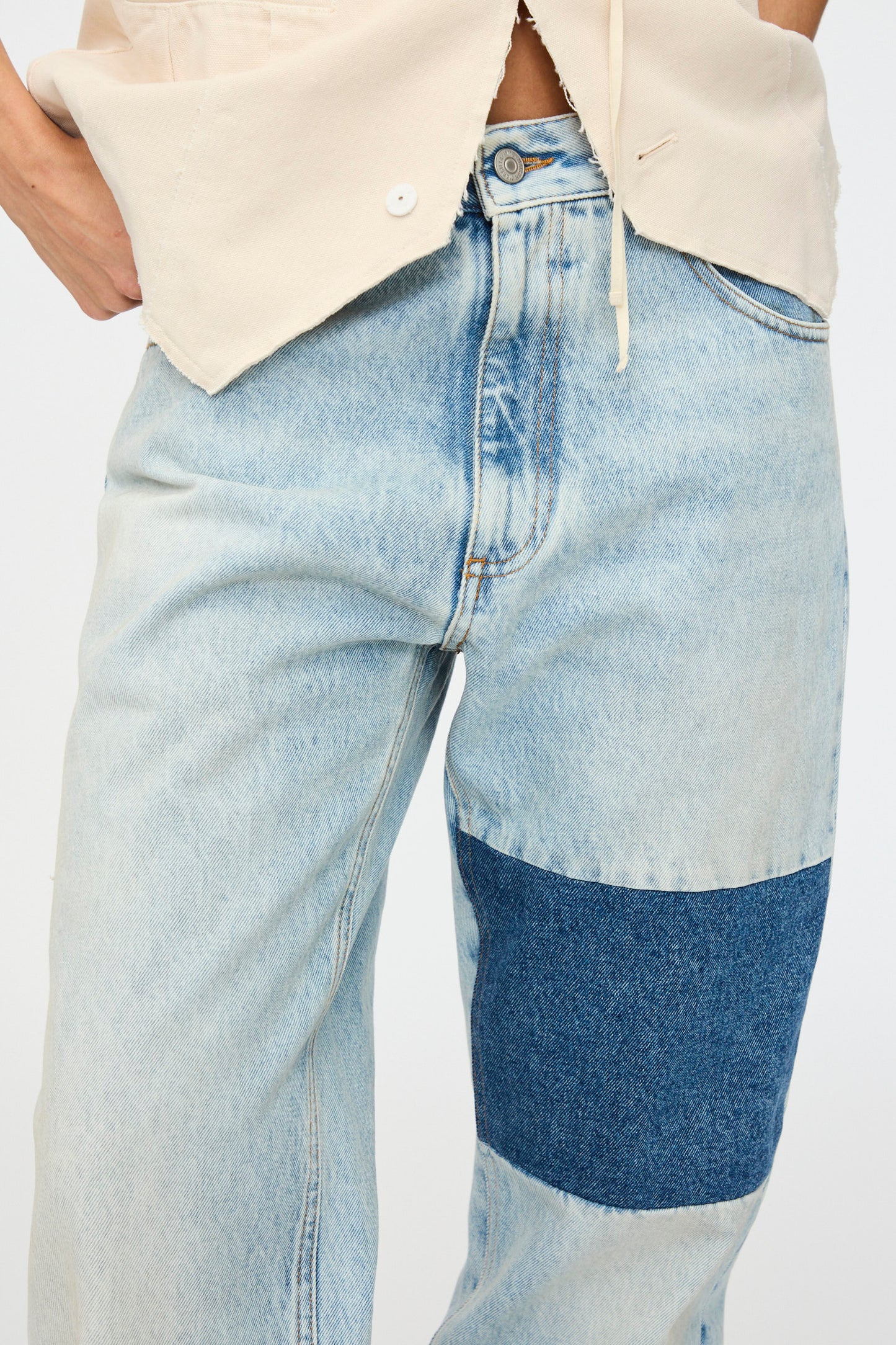 Close-up of MM6's Pants 5 Pockets, featuring light blue patchwork denim with a dark blue knee patch. A beige shirt partially covers the relaxed-fit jeans, while a hand gently rests at the hem.