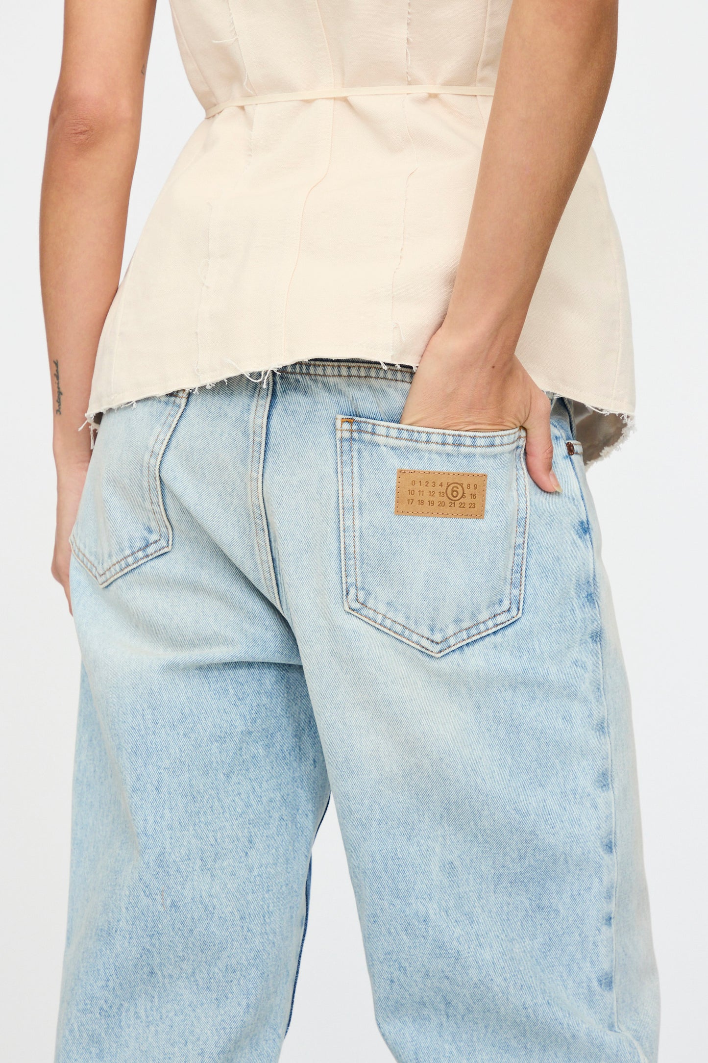 A person wearing a beige sleeveless top and MM6's Pants 5 Pockets denim jeans with a brown patch on the back pocket, hand in pocket.