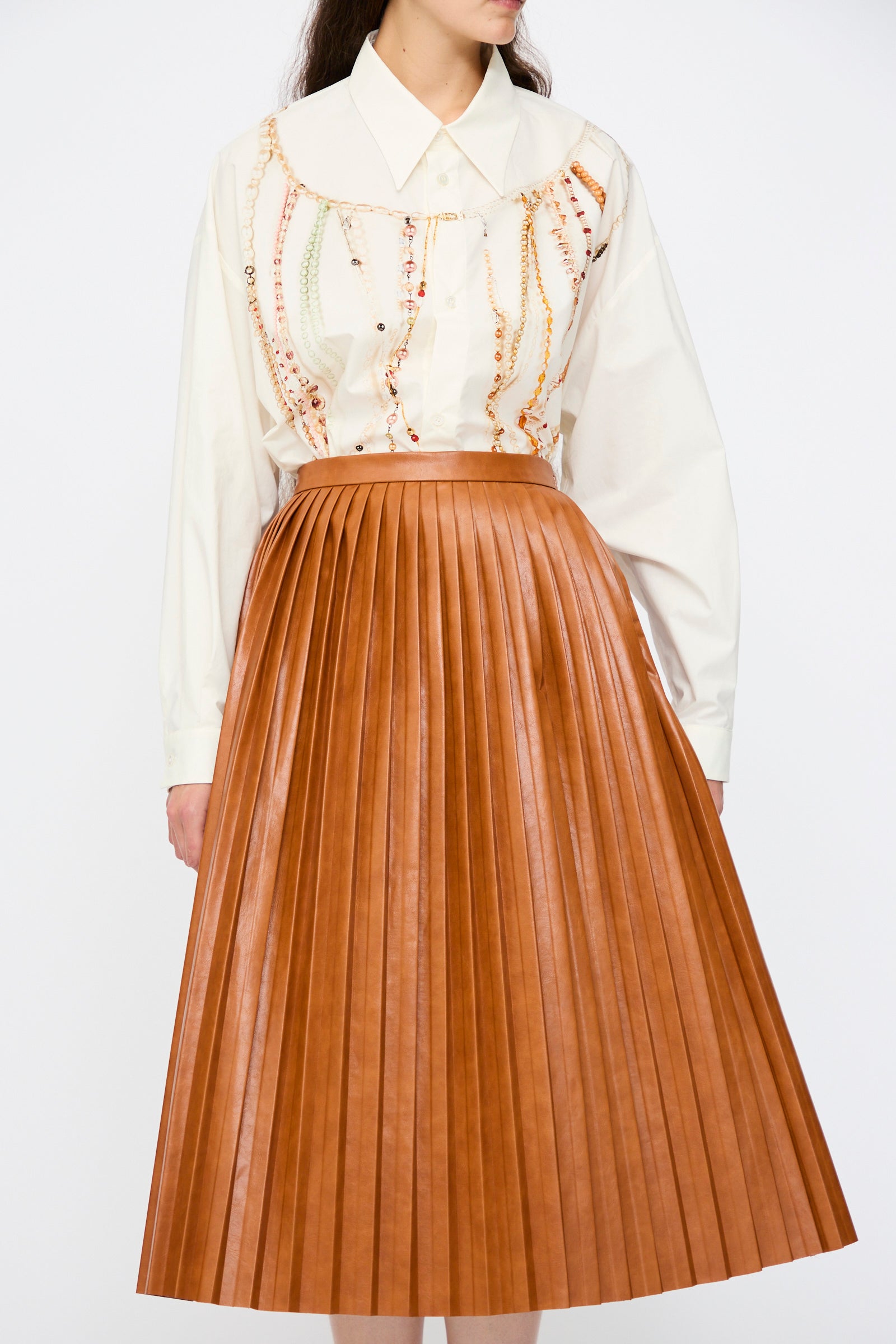 A person wearing a cream viscose chain print blouse and the MM6 Skirt in Caramel, a brown high waist pleated faux leather midi skirt, stands against a plain white background.