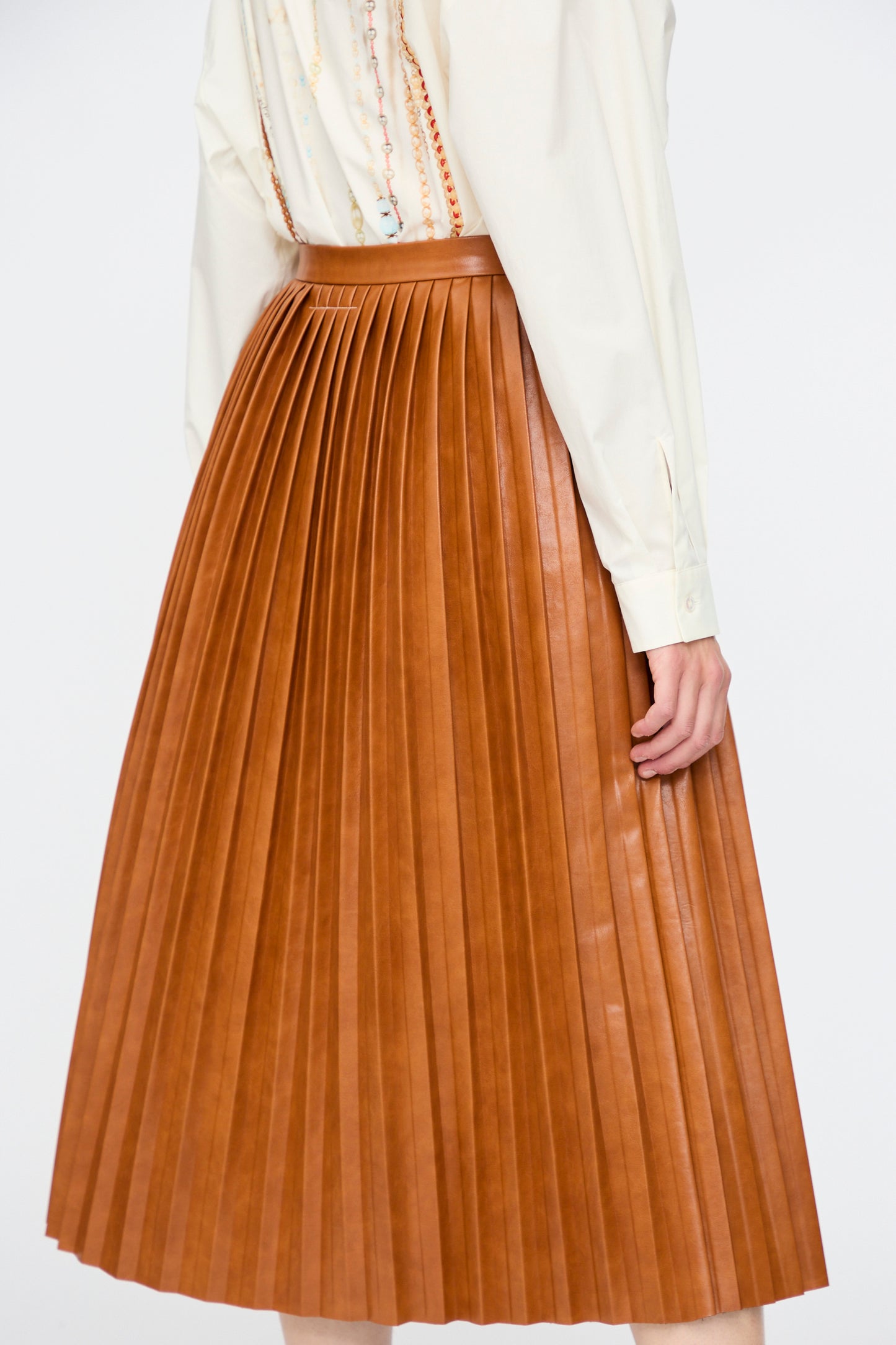 A person shown from the back wears a cream long-sleeve shirt and the MM6 Skirt in Caramel, a high-waisted pleated brown viscose skirt, against a plain background.