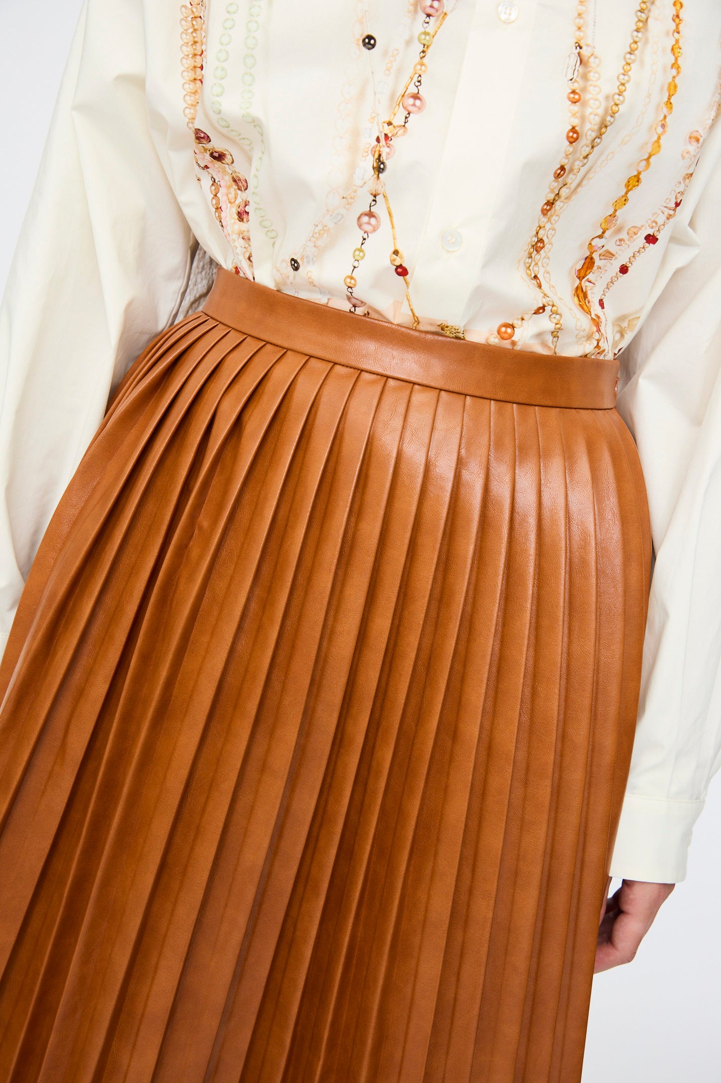 A person wearing a cream patterned viscose shirt, tucked into the MM6 Skirt in Caramel—a high-waist, pleated brown leather design.