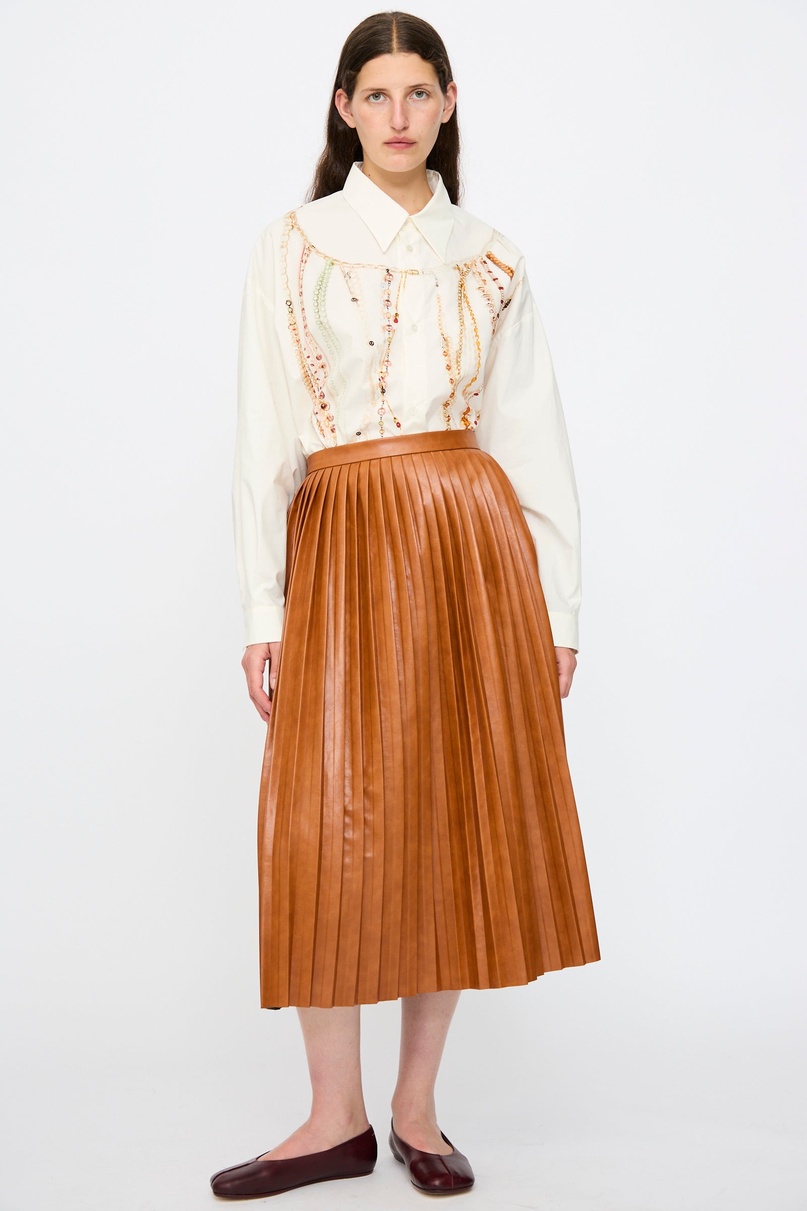 A person stands against a plain white background wearing a white blouse with embroidery, the MM6 Skirt in Caramel (high-waisted and pleated), and dark brown flat shoes.