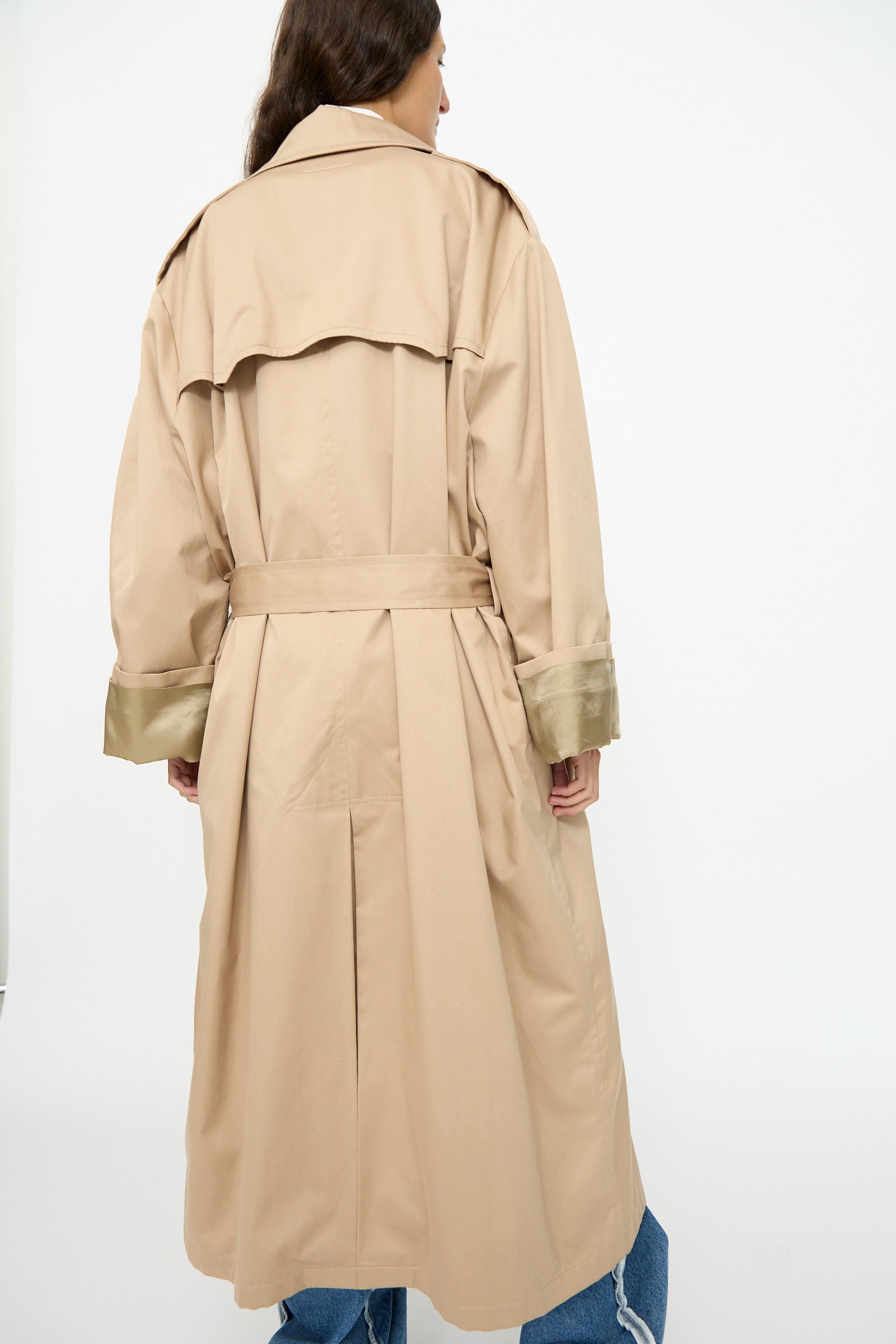 A person seen from the back wears the MM6 Trench Coat in Sand, a long, double-breasted Italian-made coat with a belt and wide cuffs, styled with blue jeans.