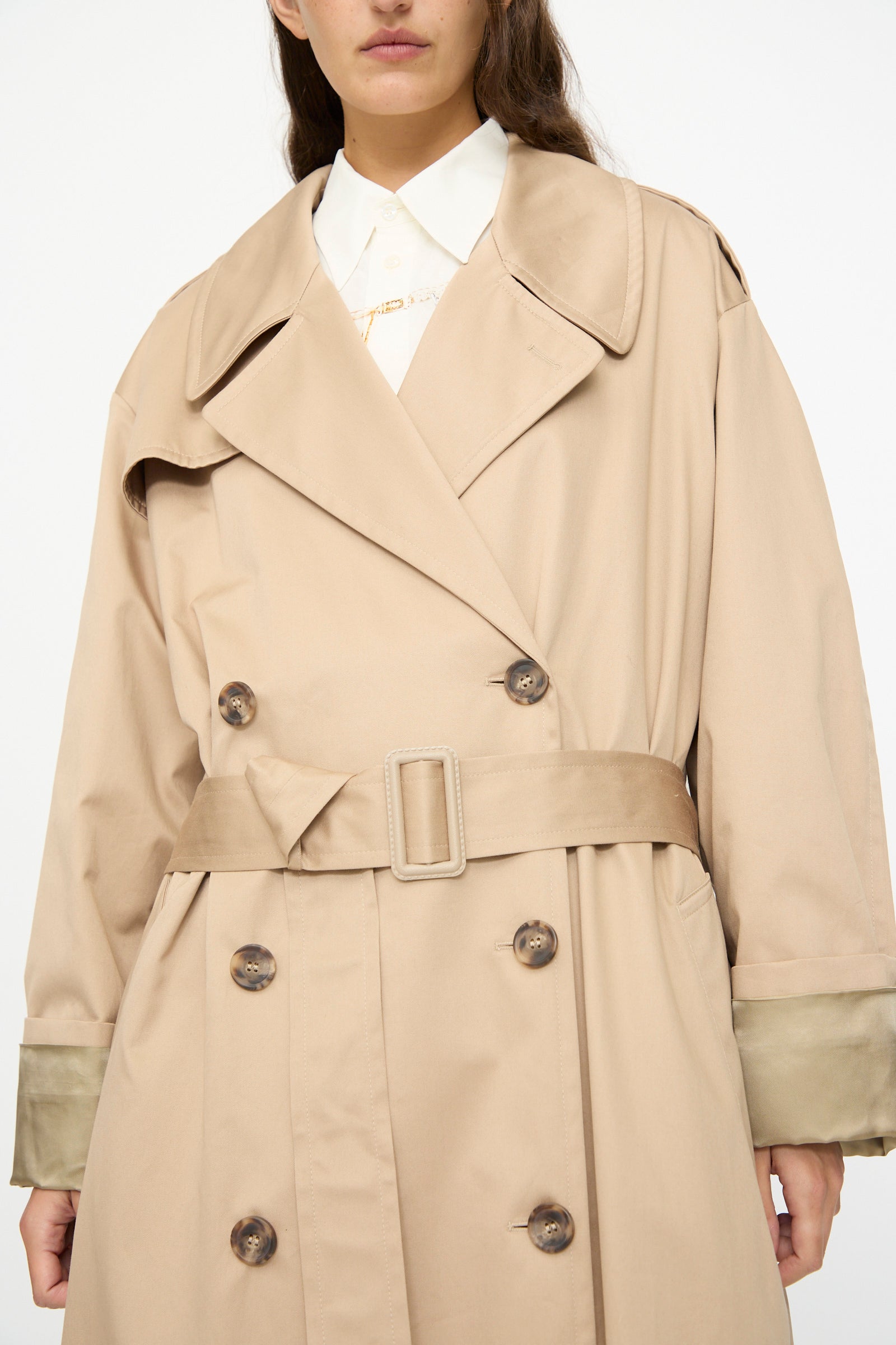 A person wearing the MM6 Trench Coat in Sand, a double-breasted, belted coat made in Italy, over a white collared shirt, shown from shoulders to knees.