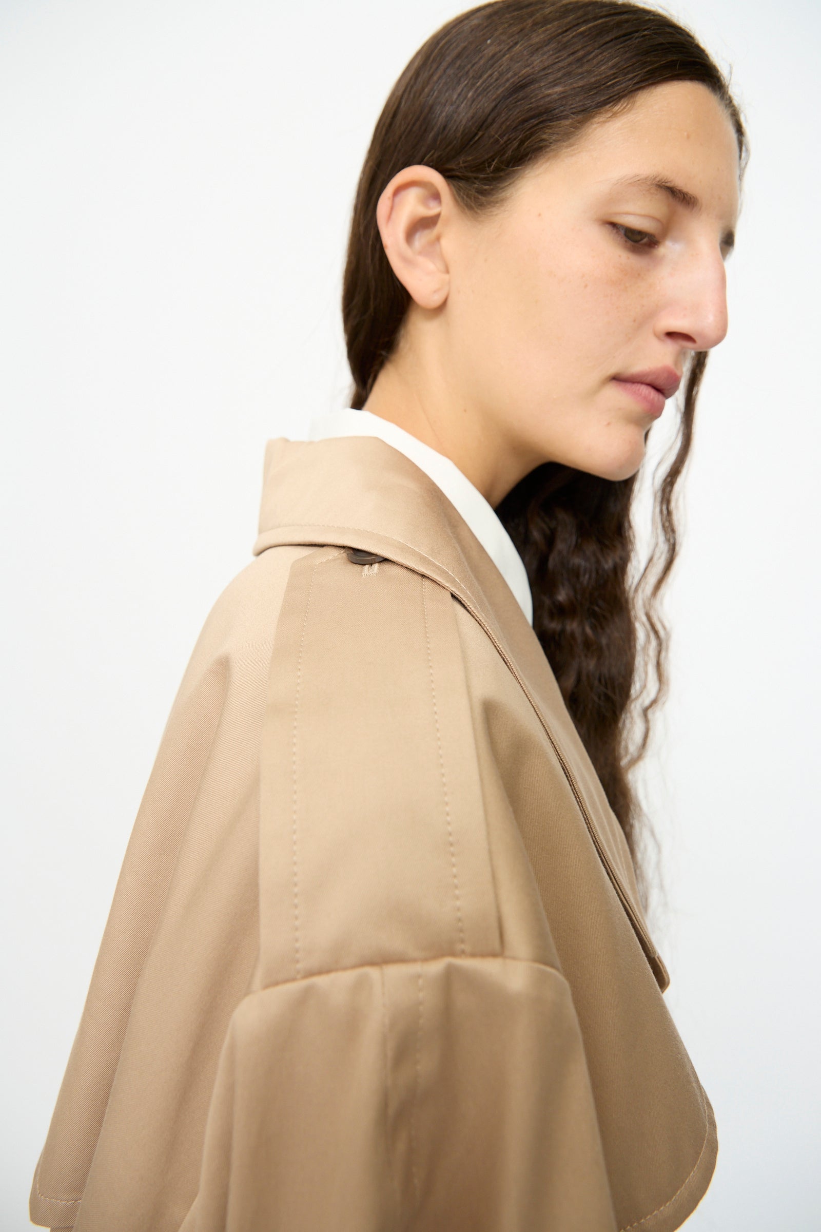 A person with long brown hair wears the MM6 Trench Coat in Sand over a white shirt, standing against a plain light background.