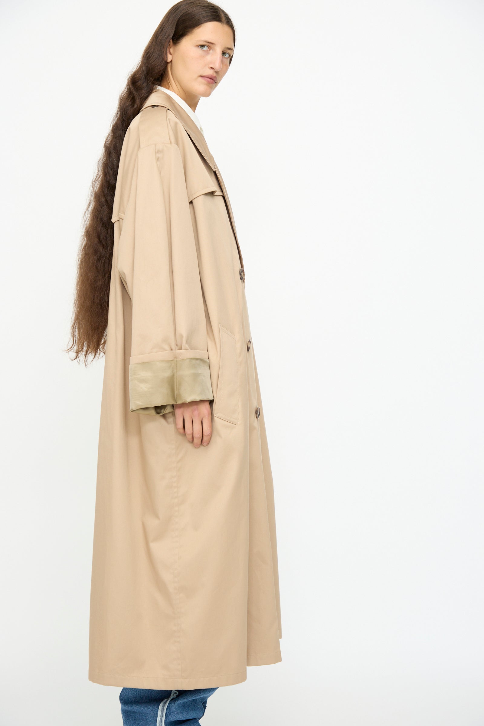 A person with long brown hair wears the MM6 Trench Coat in Sand with wide sleeves and blue jeans, standing sideways against a plain white background.