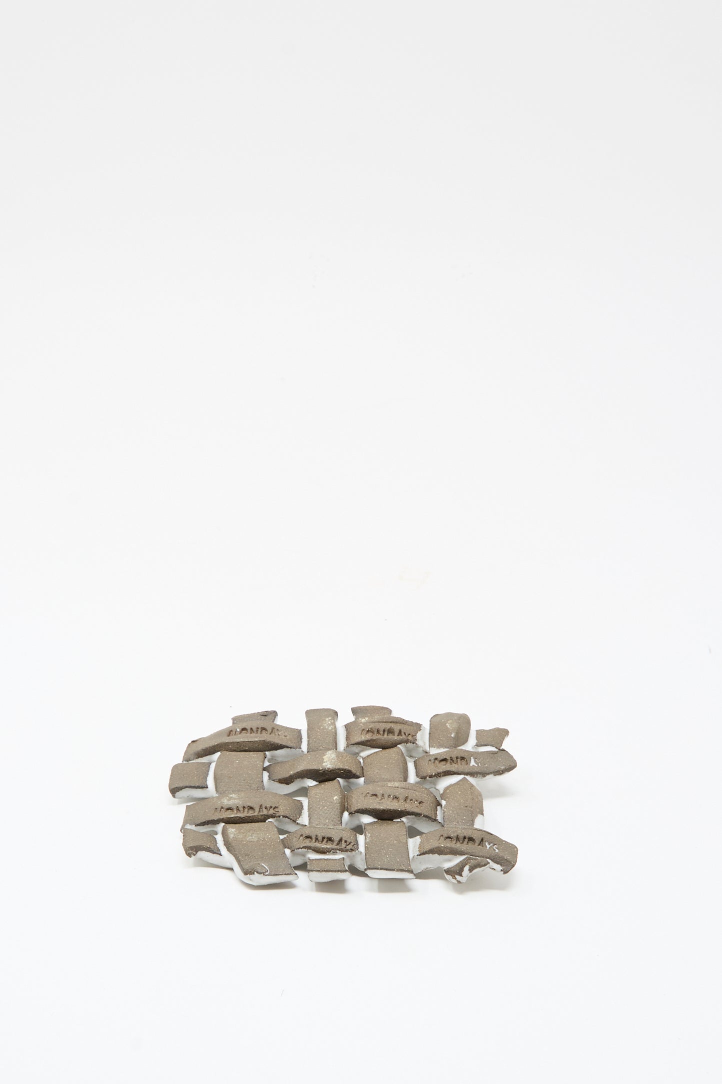A small pile of irregular metallic pieces with text rests beside the MONDAYS Woven Dish in White, a ceramic piece finished with a white glaze, all arranged on a plain white background.
