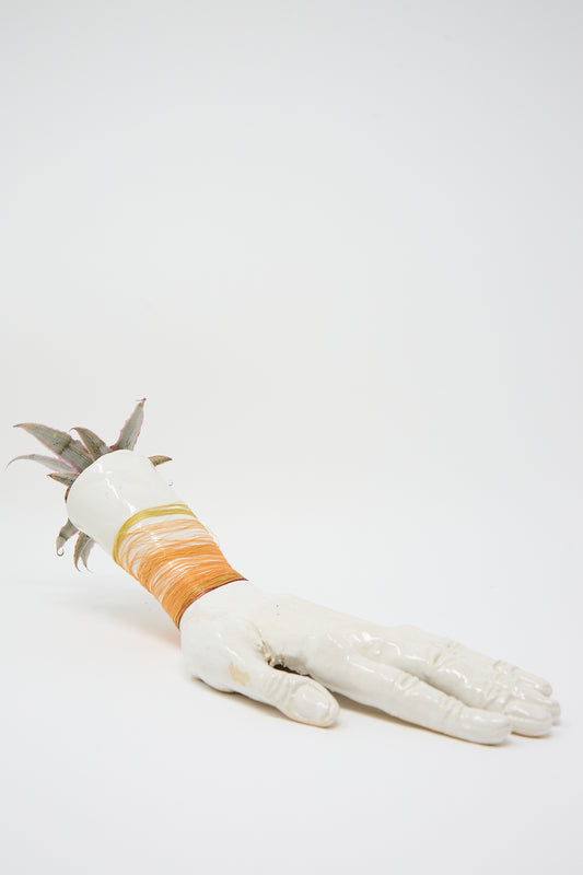 A ceramic sculpture of a hand lies flat, serving as a unique Ceramic Hand Sculpture with Orange Thread. The wrist is wrapped with orange thread detail, and a small plant sprouts from the end of the wrist. This piece is handmade in Brooklyn by Monty J.