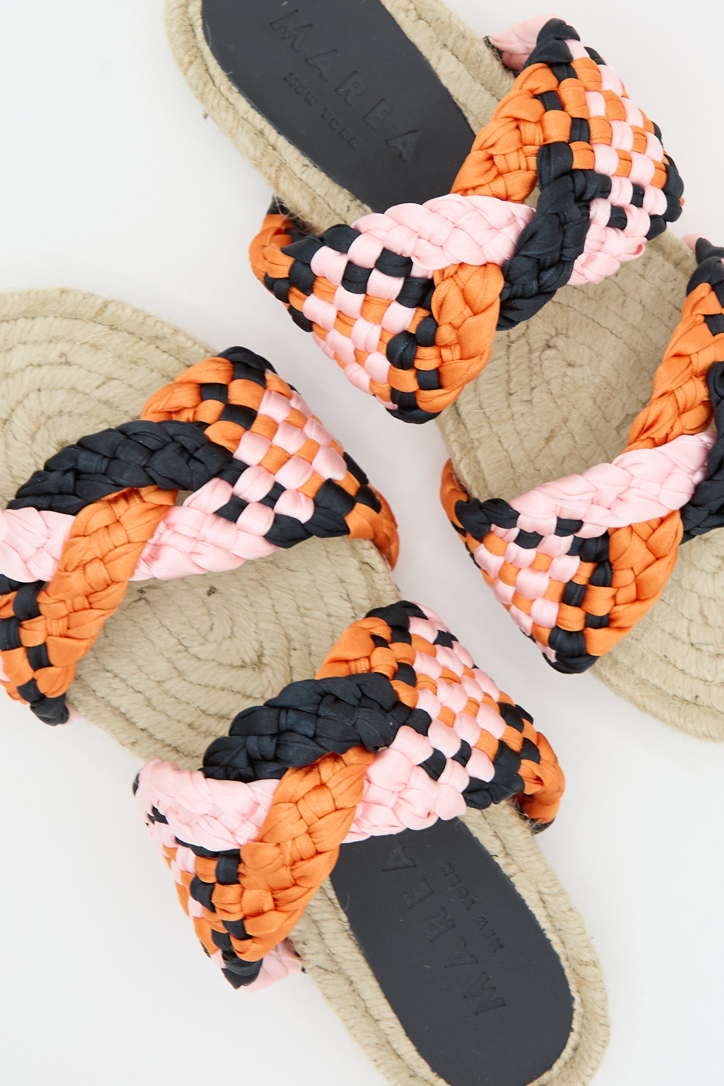 Top view of two pairs of Marea Braided Satin Espadrille sandals with orange, pink, and black straps on a beige background.