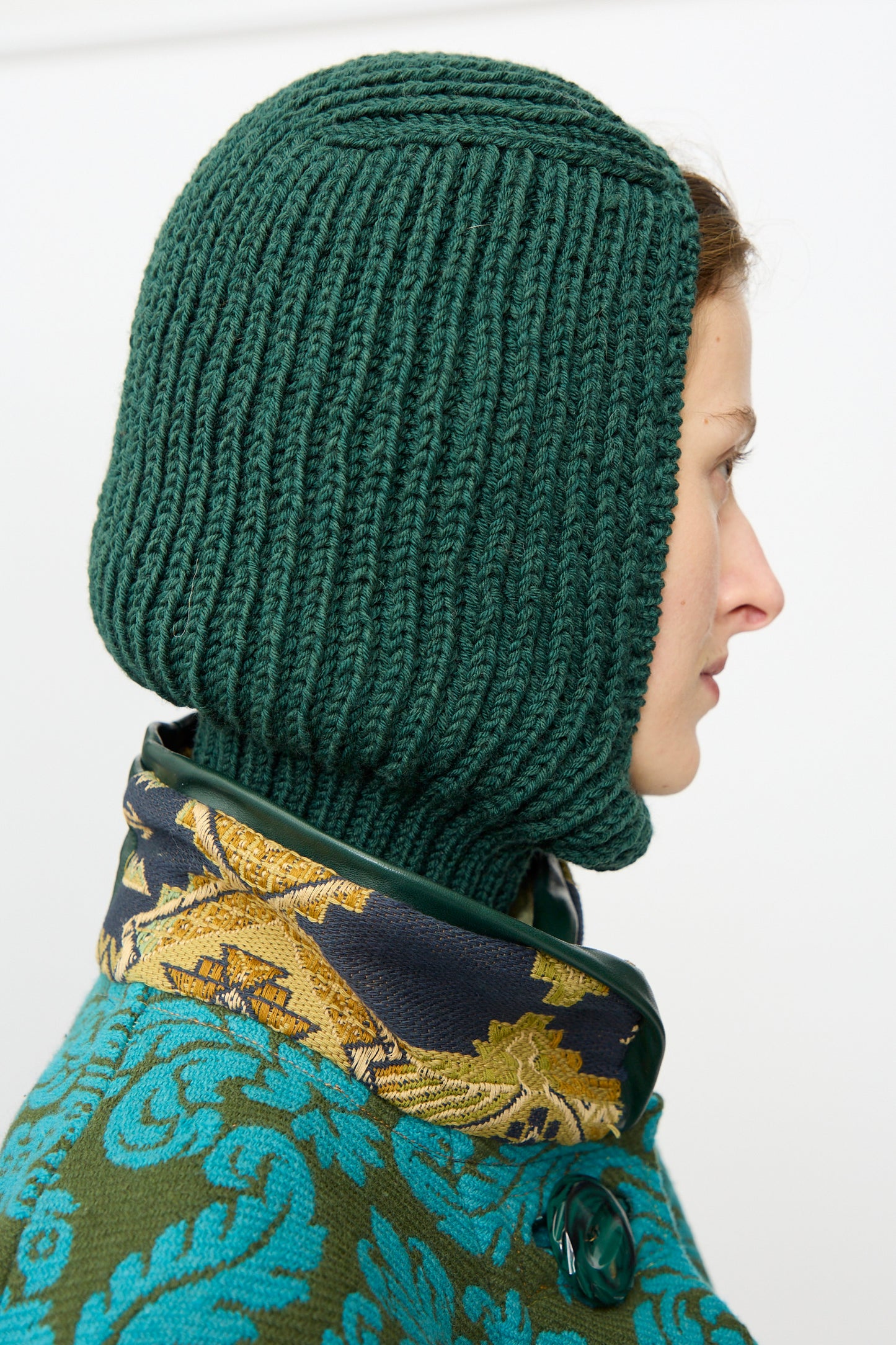 A person in profile wears the Maria La Rosa Wool Balaclava Lina in Emerald and a coat with a blue and gold pattern featuring a large green button.