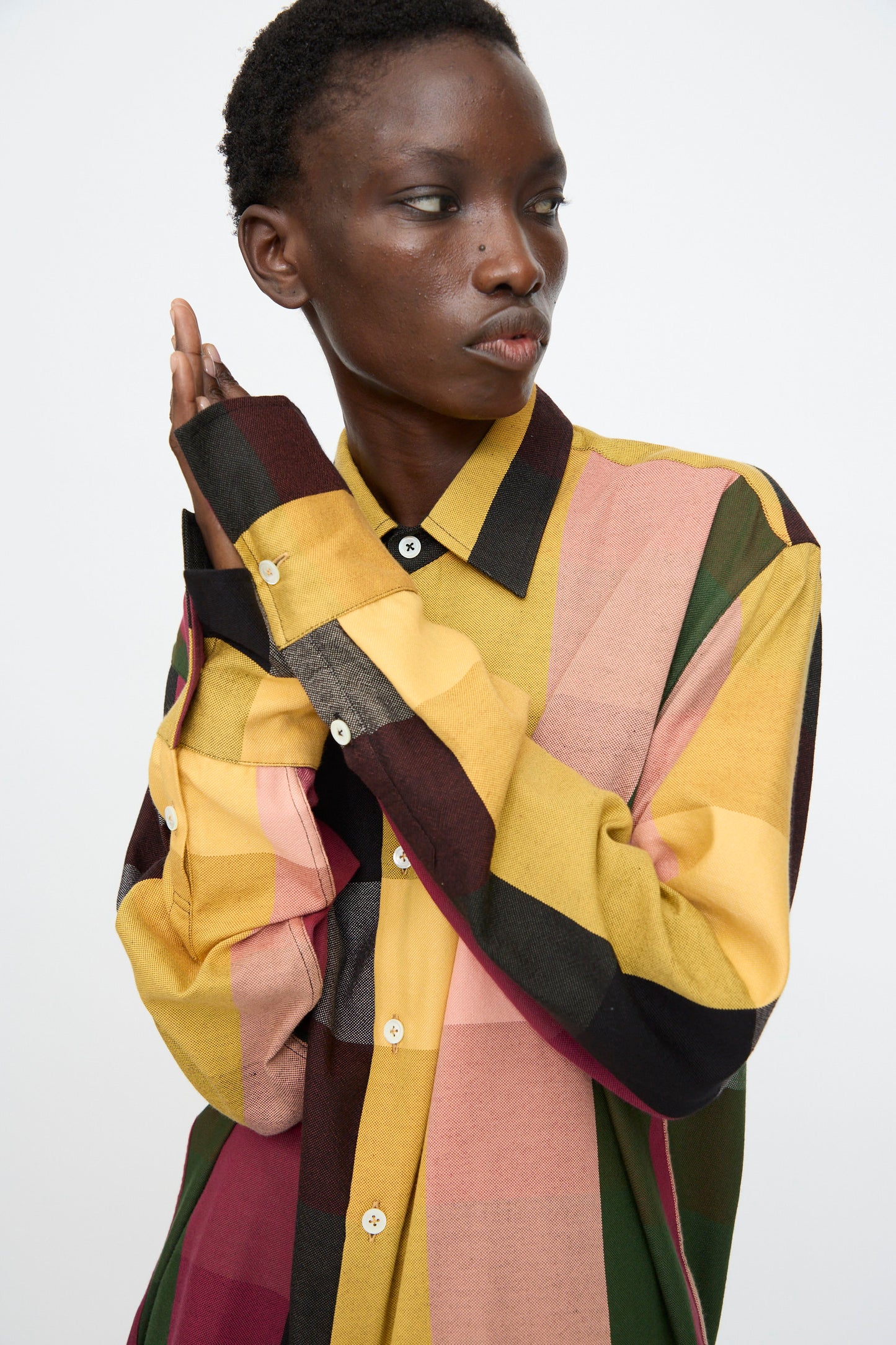 A person wearing the Marrakshi Life Button Down Shirt in Plaid Multi, a vibrant cotton blend, looks to the side with hands near their face against a plain background.