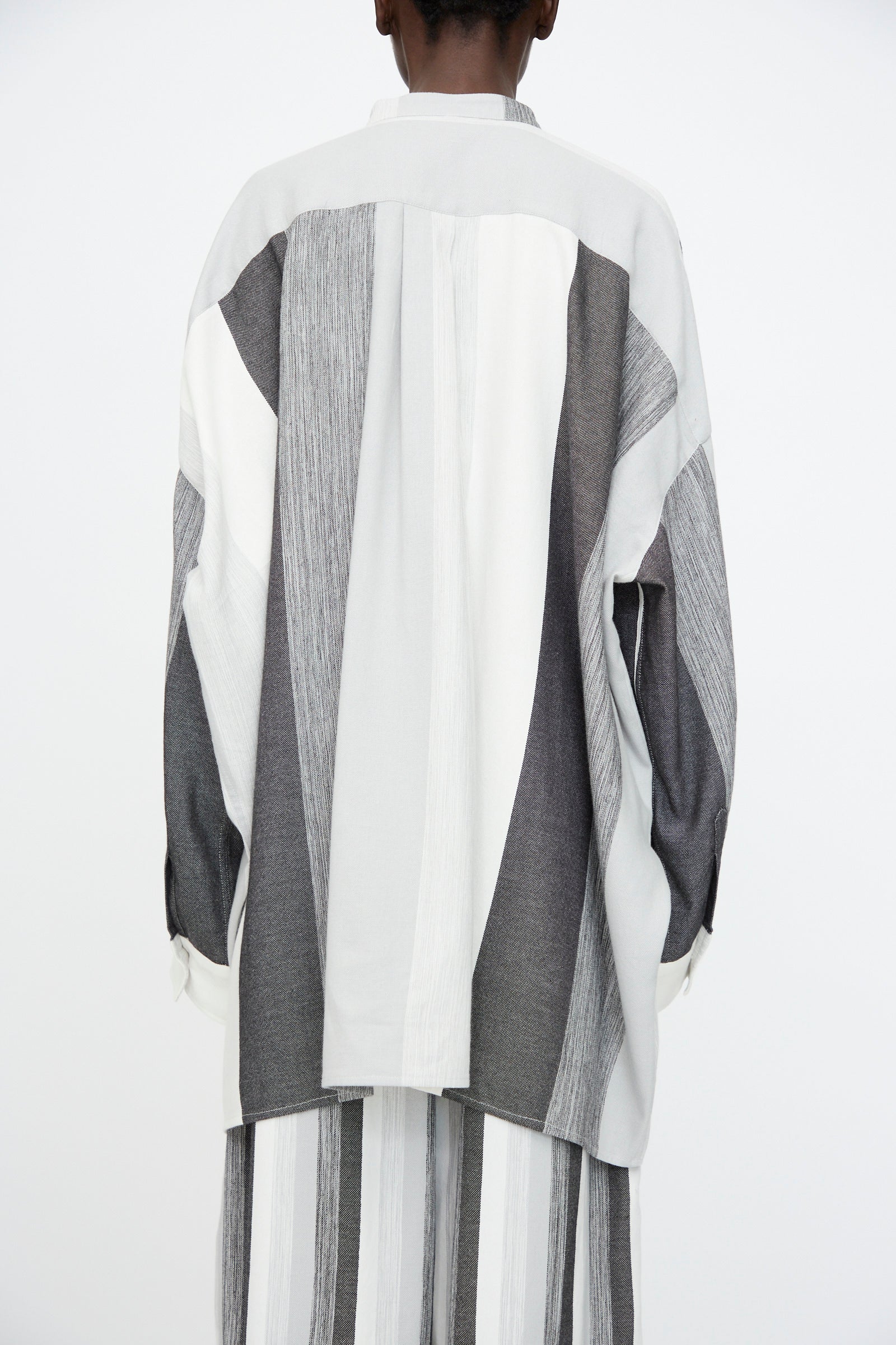 A person wearing the Marrakshi Life Nero Collar Oversized Shirt in Grey Stripe, featuring vertical and diagonal gray and white panels, paired with matching striped pants, shown from the back against a plain background.