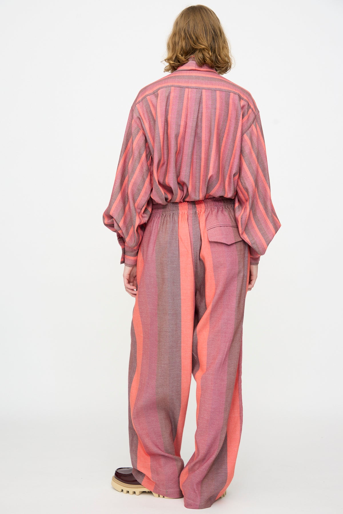 A person stands with their back to the camera, wearing Marrakshi Life’s The Parachute Pant in Stripe Multi—a loose-fitting, pink and coral handwoven cotton pant paired with a matching striped shirt and beige-soled shoes, set against a plain white background.