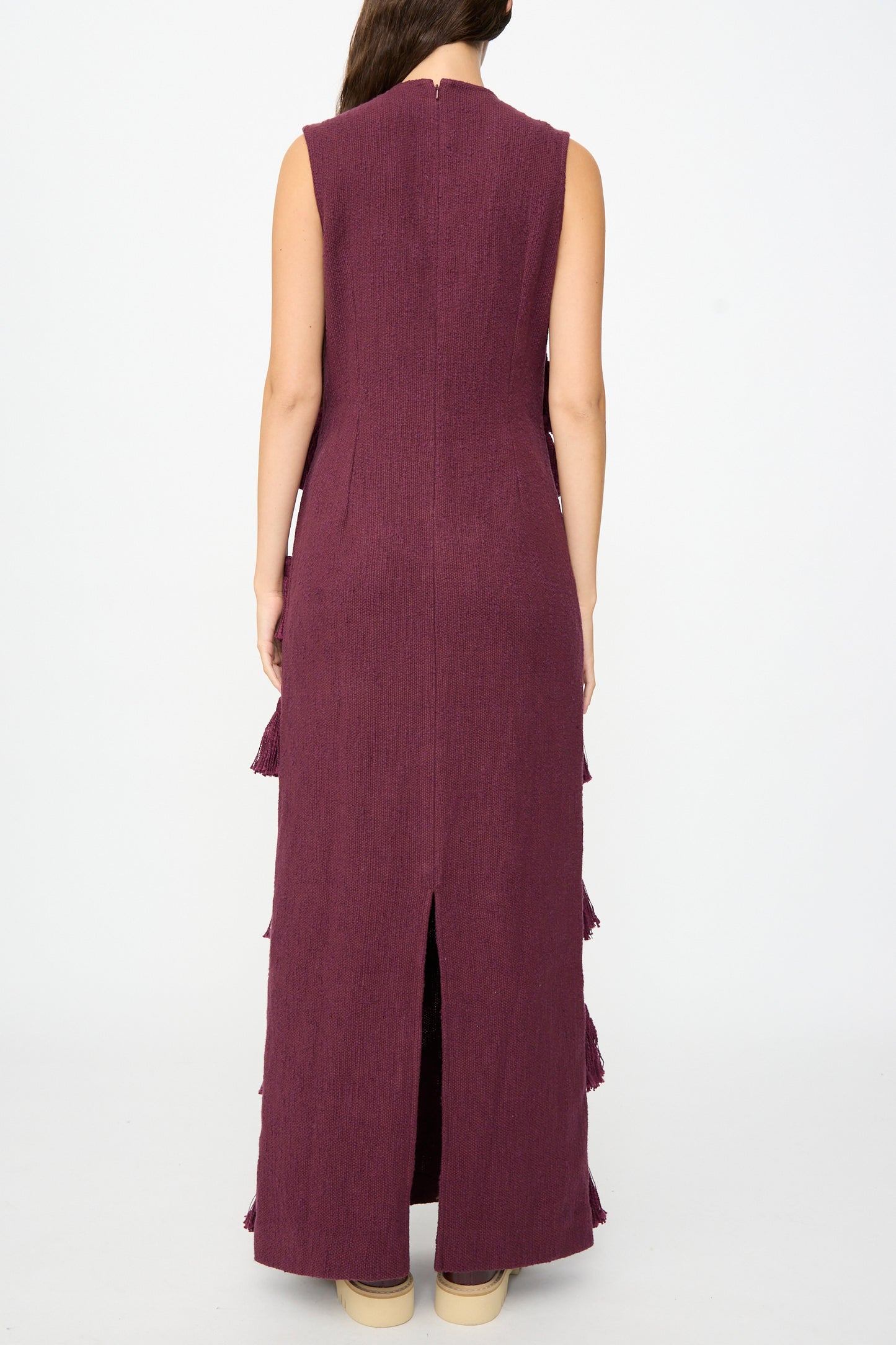 A person stands facing away, wearing Marrakshi Life’s The Pompom Dress in Merlot Flamme—a sleeveless maxi crafted from textured, handwoven cotton in purple with a back slit—paired with beige shoes. This unique piece is handmade in Morocco.
