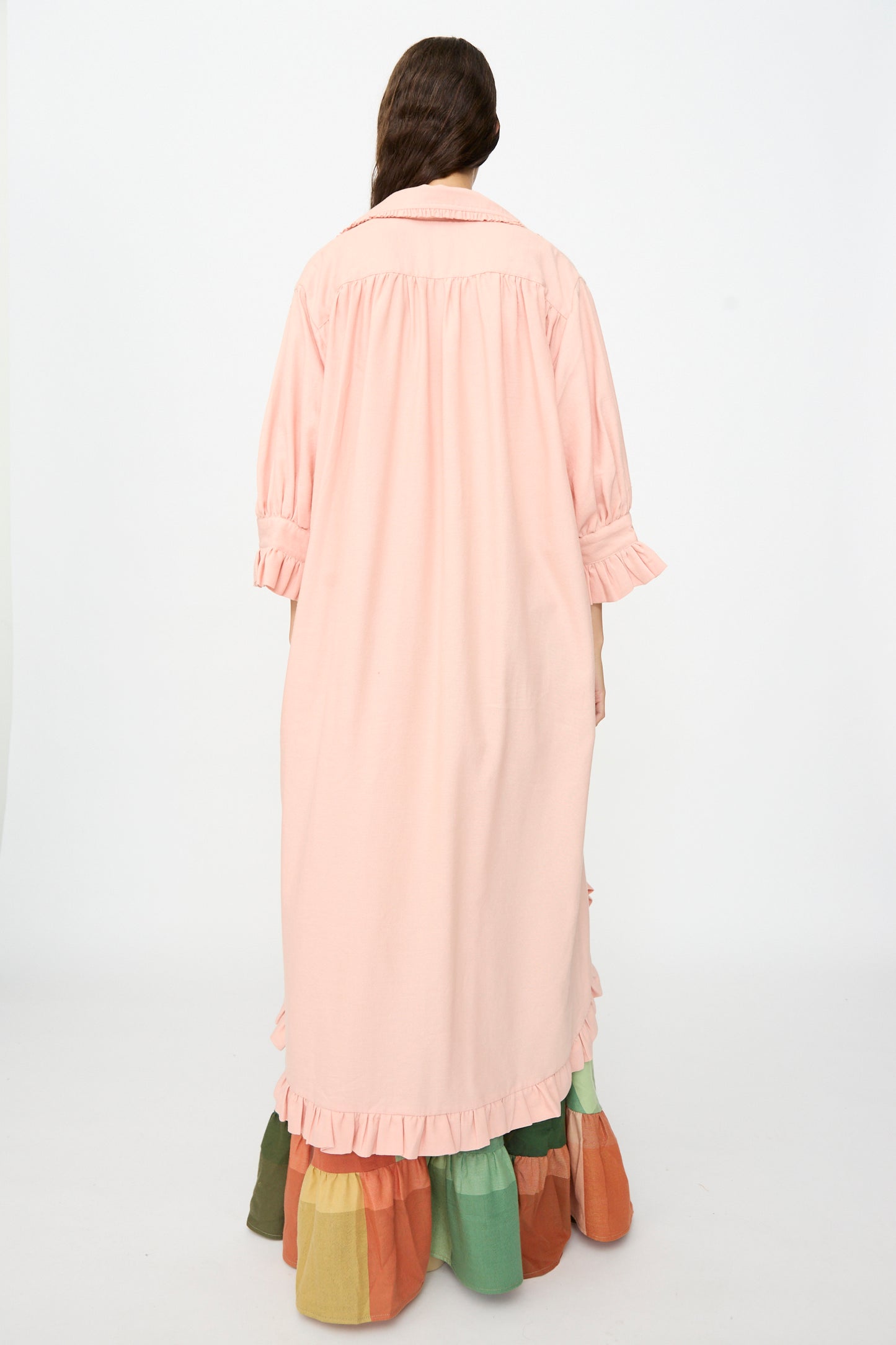 A person with long brown hair stands facing away, wearing Marrakshi Life’s The Ruffled Tunic in Flesh—an oversized, handwoven cotton dress handmade in Morocco, featuring ruffled cuffs and hem. The tunic is styled over a multicolored tiered skirt against a plain white background.