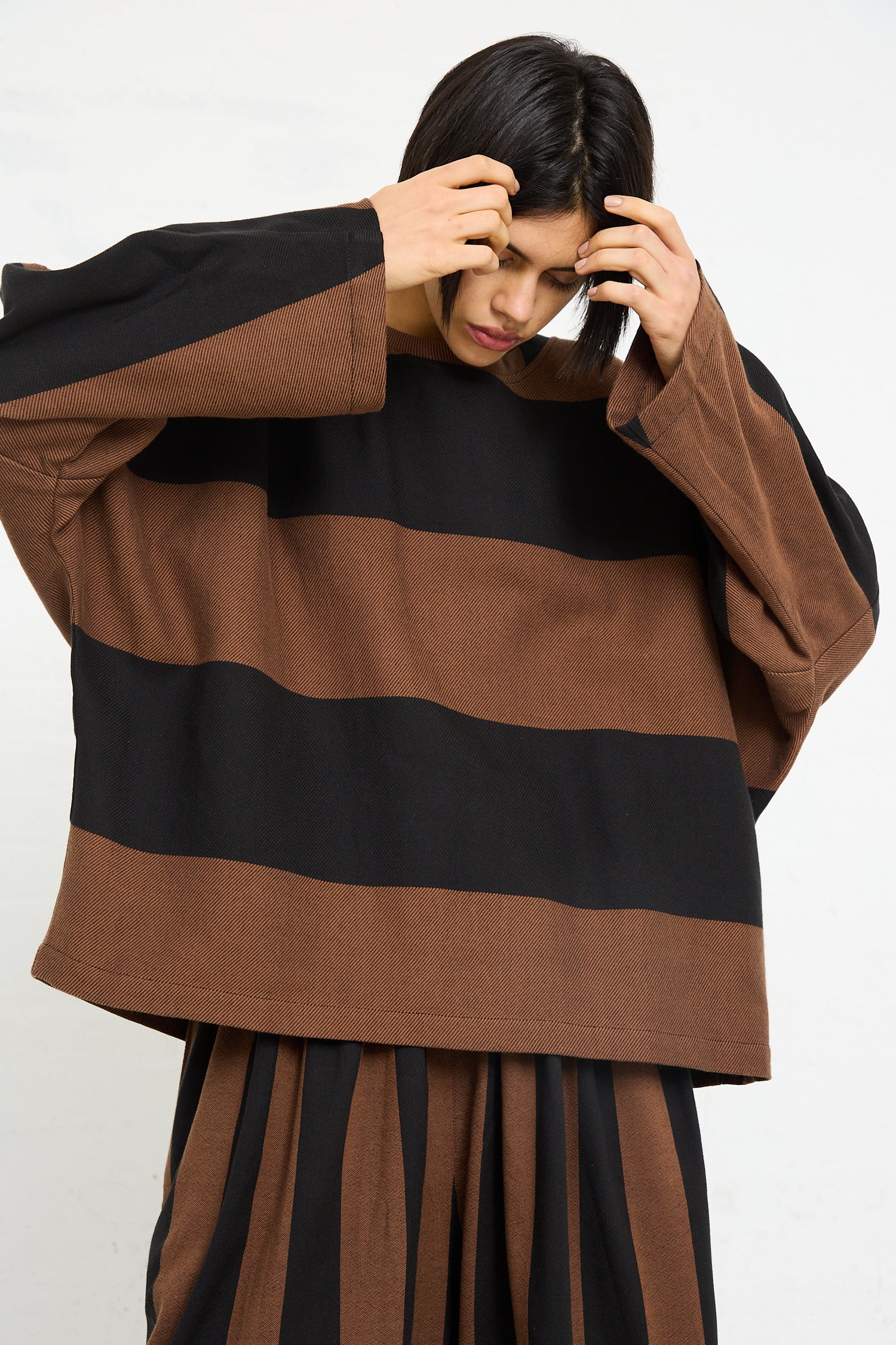 Woman in a striped Marrakshi Life Twill Sweater in Black and Brown Stripe with oversized long sleeves, holding her head, appearing thoughtful or concentrated.