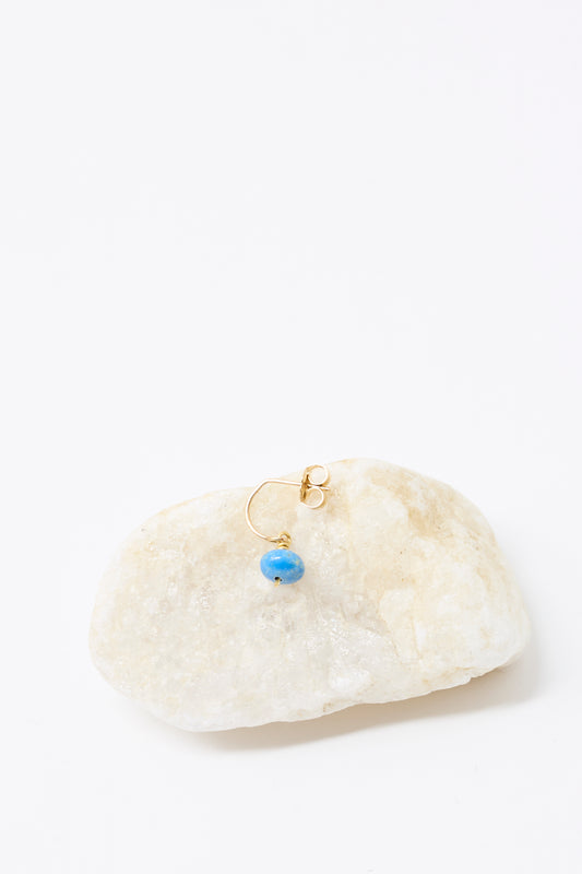 The Mary MacGill 14K Crescent Charm Single Earring in Ceruleite features a small blue bead and is shown on light-colored stone against a white background.