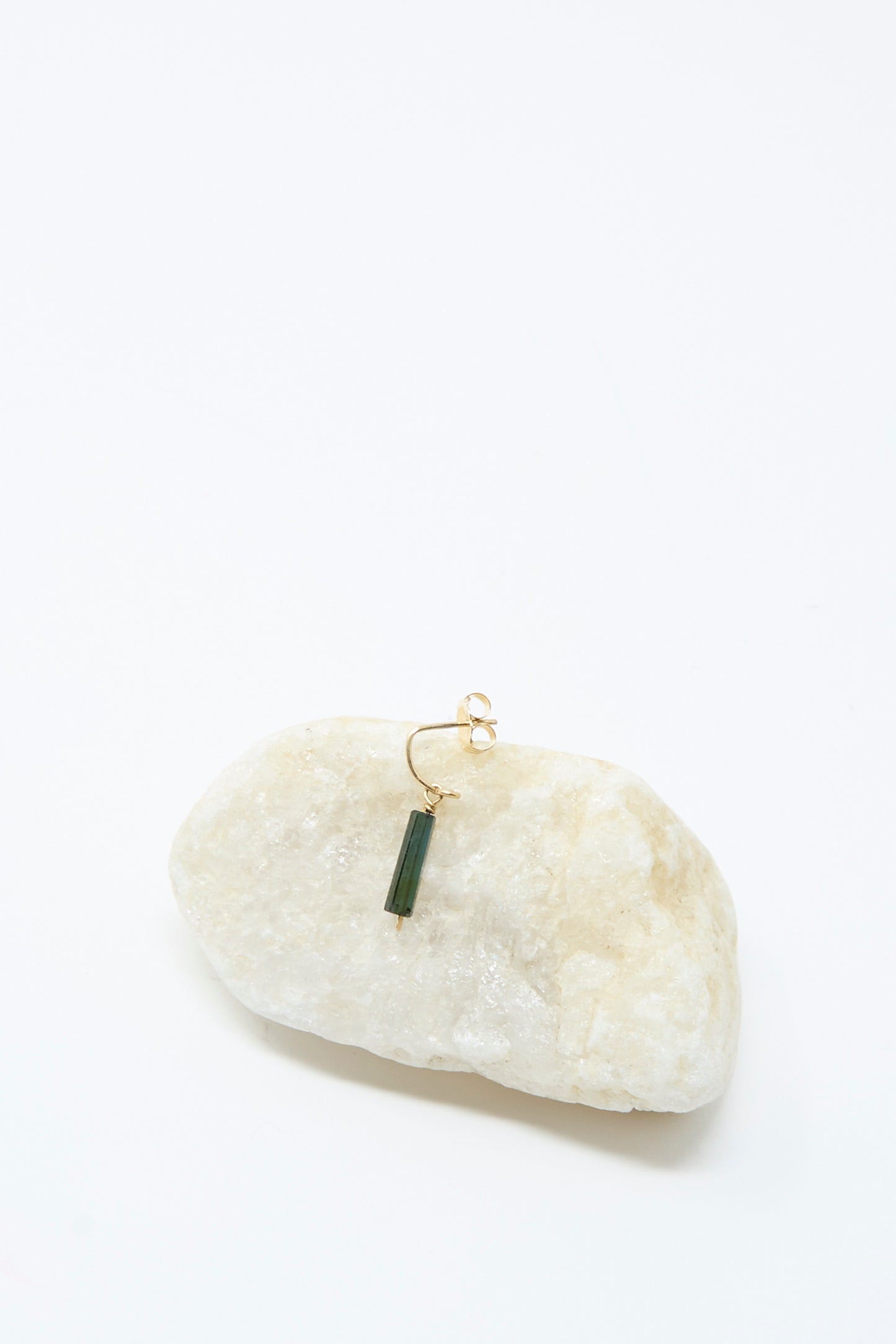 The Mary MacGill 14K Crescent Charm Single Earring in Dark Tourmaline Tube, featuring a handmade vertical green stone, is displayed on a smooth light-colored rock against a white background.