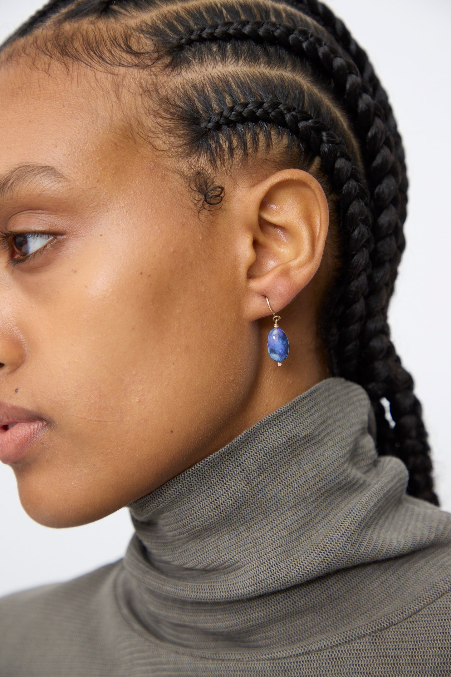 Close-up of a person with braided hair in a gray turtleneck, facing left, wearing the Mary MacGill 14K Crescent Charm Single Earring in Dumortierite Quartz.