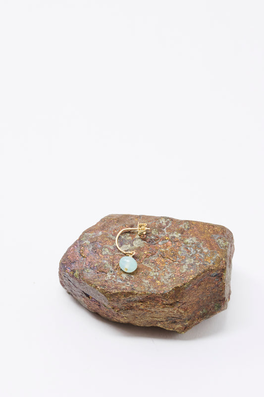 The Mary MacGill 14K Crescent Charm Single Earring in Mint Opal is shown atop a rough brown rock, set against a white background.
