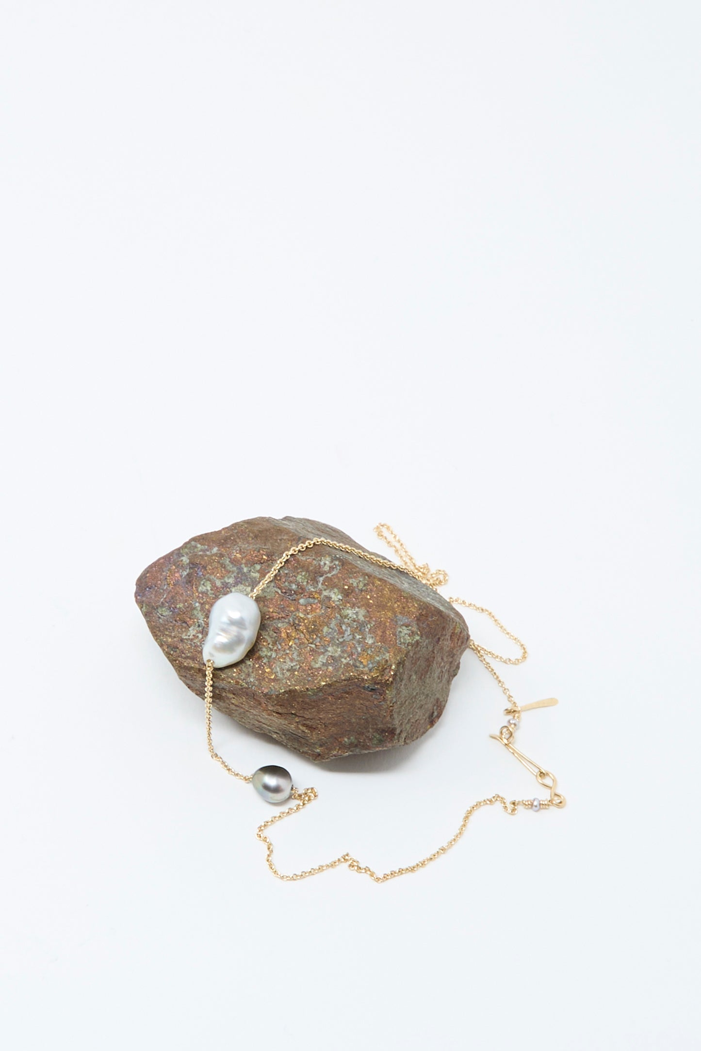 The Mary MacGill 14K Necklace featuring White Keshi and Silver Tahitian Pearls is draped over a rough brown stone against a plain white background.