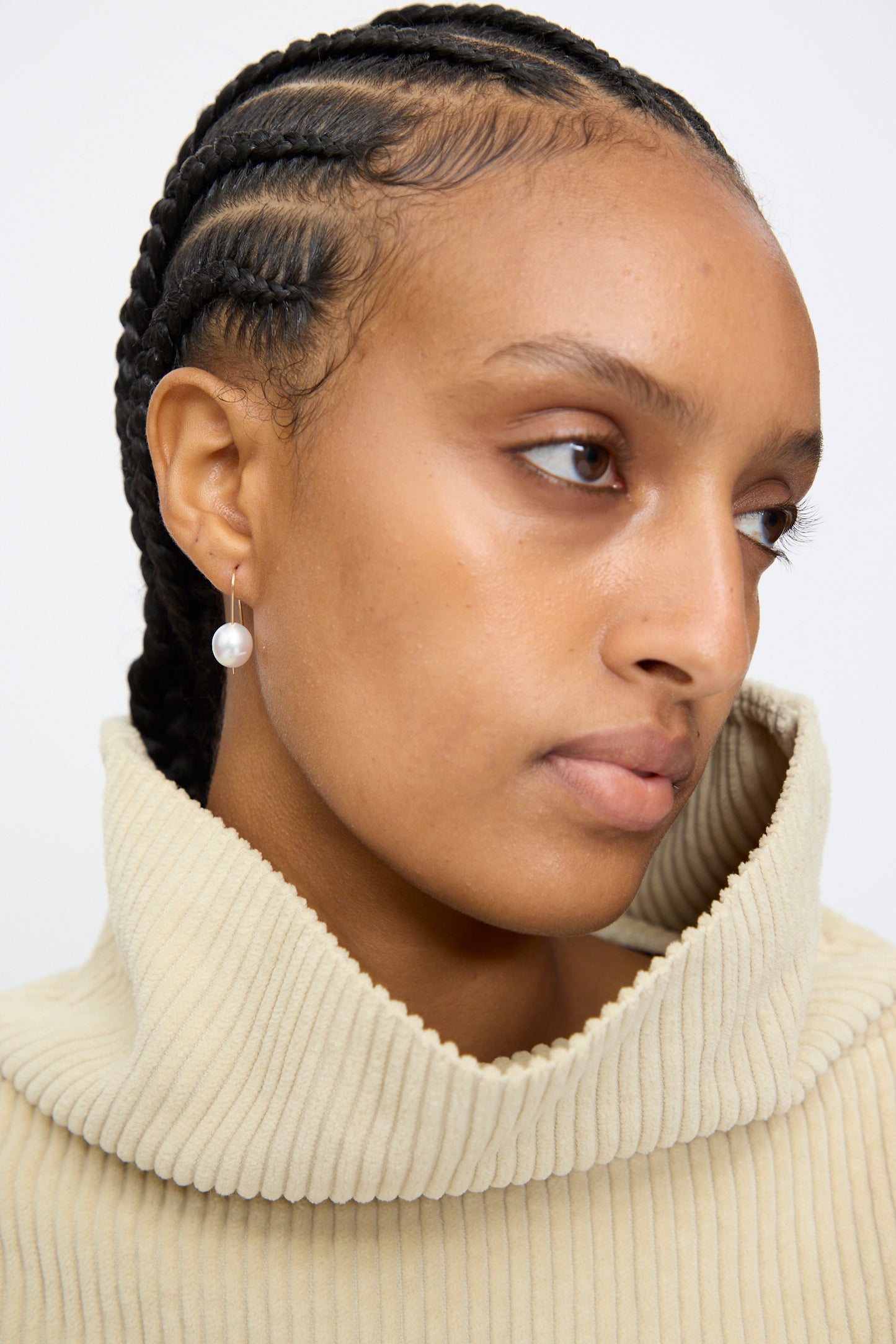 A person with cornrow braids, wearing a beige ribbed turtleneck and a single Mary MacGill 14K Tahitian Pearl Drop Earring in White, looks slightly to the side.