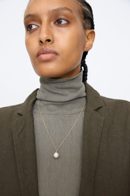 A person in a dark olive jacket and taupe turtleneck wears the Mary MacGill 14K Tahitian Pearl Drop Necklace in White, featuring a single pearl pendant, while looking slightly to the side against a plain background.
