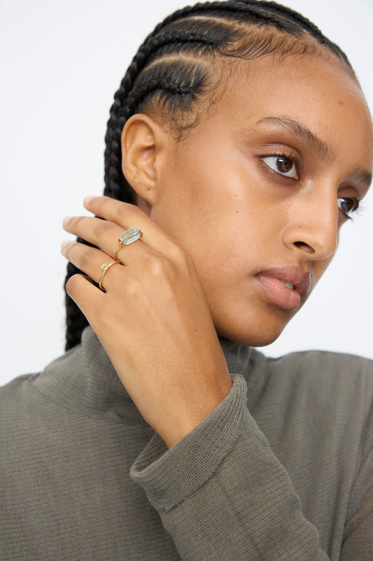 A person with braided hair, in a gray turtleneck and multiple gold rings—including Mary MacGill’s 14K Ring in Aquamarine Tube with a large rectangular gemstone—poses with their hand near their face.