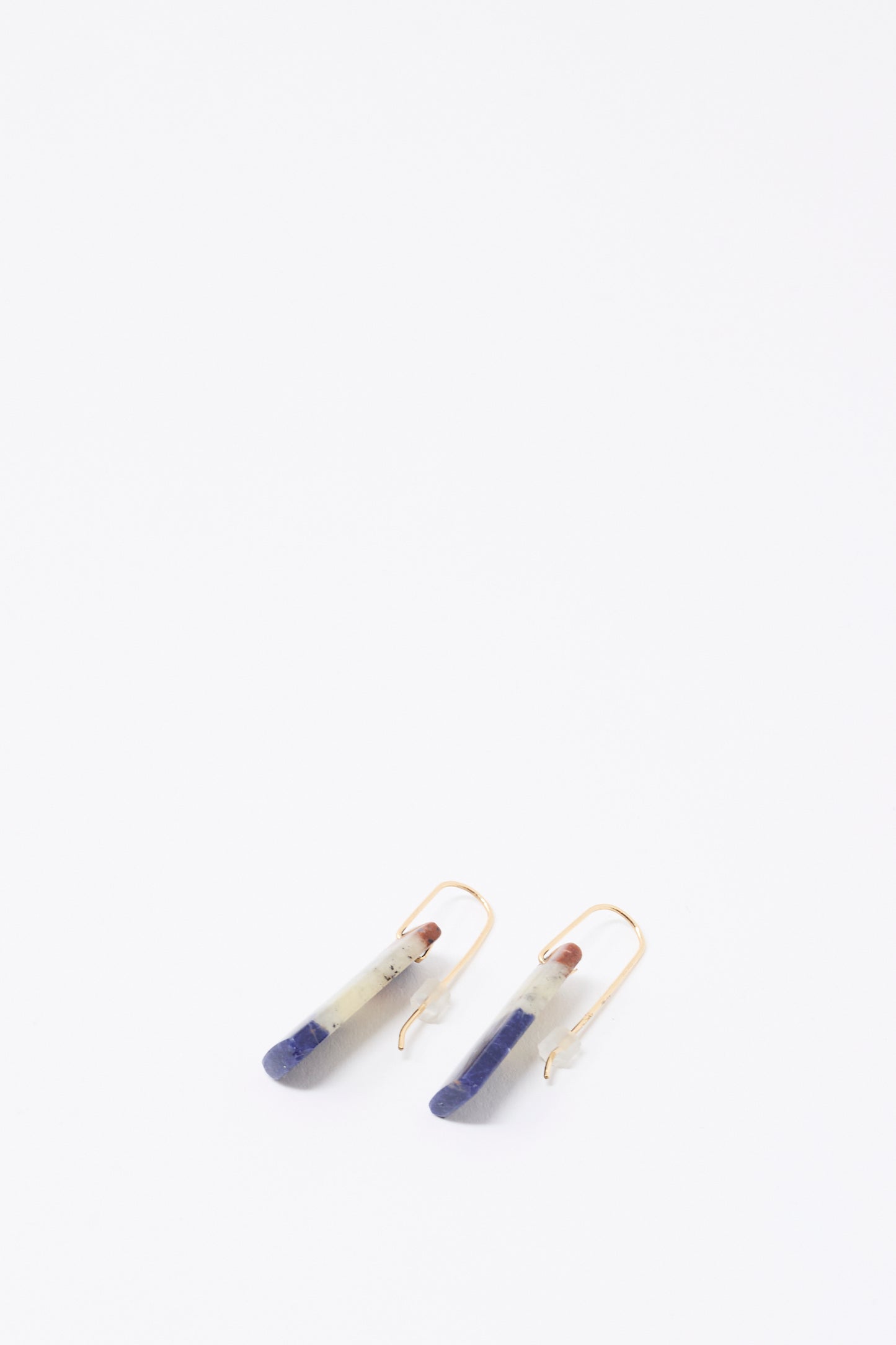 Mary MacGill’s Stone Drop Earrings in Sodalite feature handmade, elongated blue and gray stone pendants with gold hooks, shown on a white background.