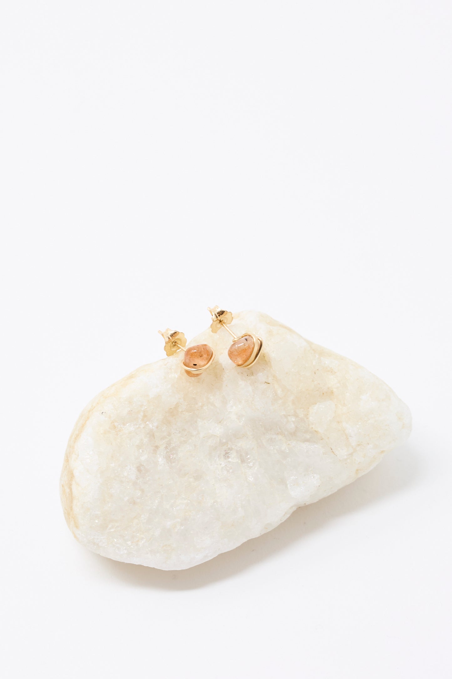 The Stone Studs in Sunstone by Mary MacGill are small gold-filled earrings featuring oval sunstone gems, shown atop a white textured rock with a clean white background.