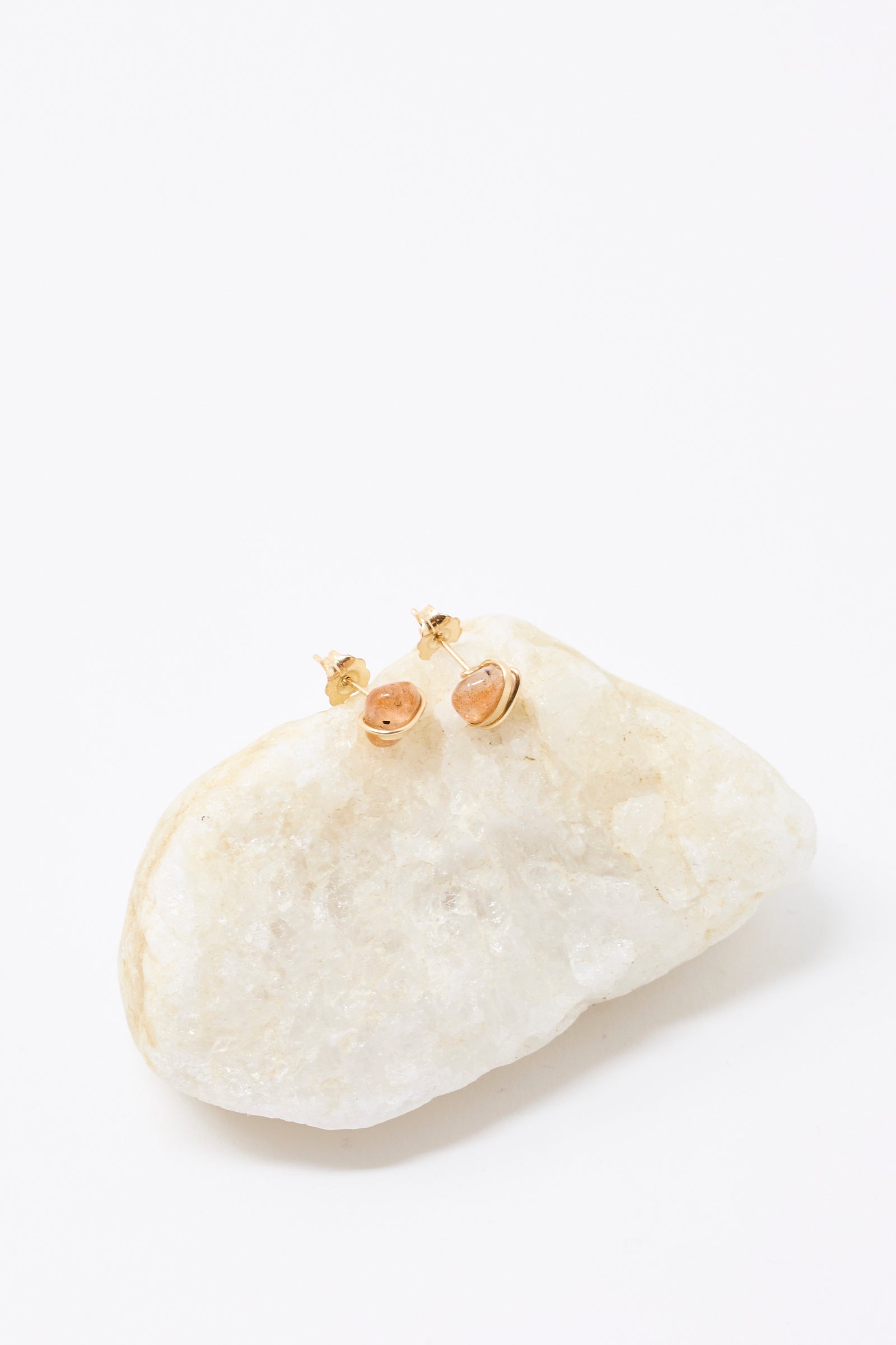 The Stone Studs in Sunstone by Mary MacGill are small gold-filled earrings featuring oval sunstone gems, shown atop a white textured rock with a clean white background.