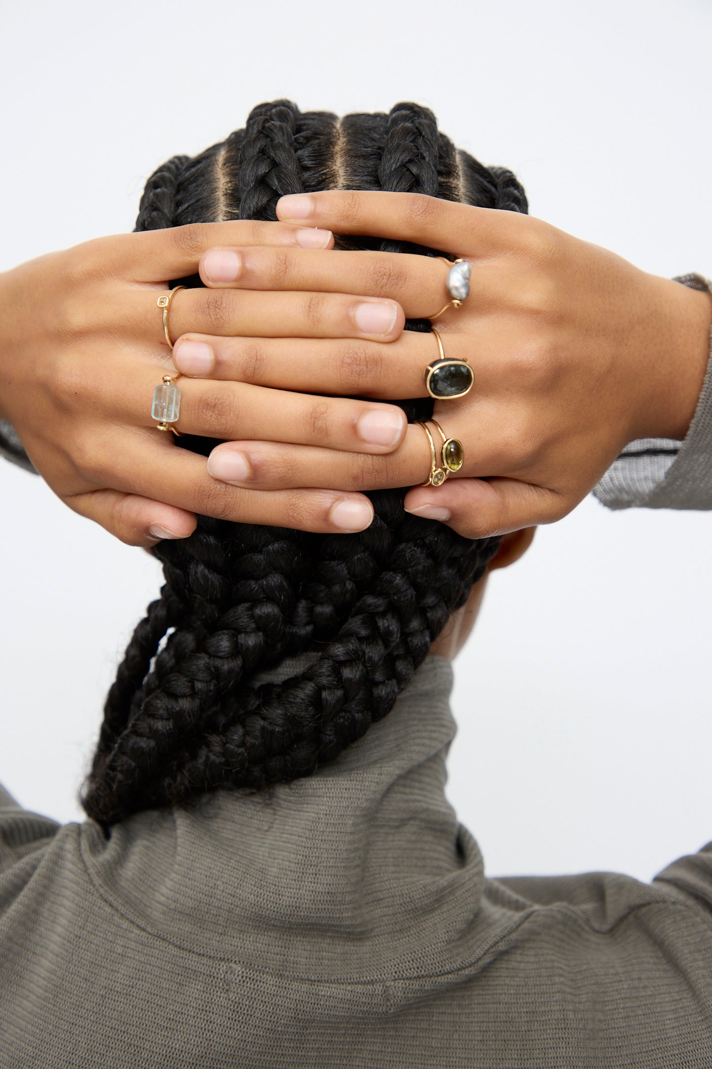 A person with cornrow braids, wearing a gray turtleneck, clasps their hands behind their head to show off several gold rings—most notably Mary MacGill’s 14K Floating Ring in Green Tourmaline—each set with unique stones.