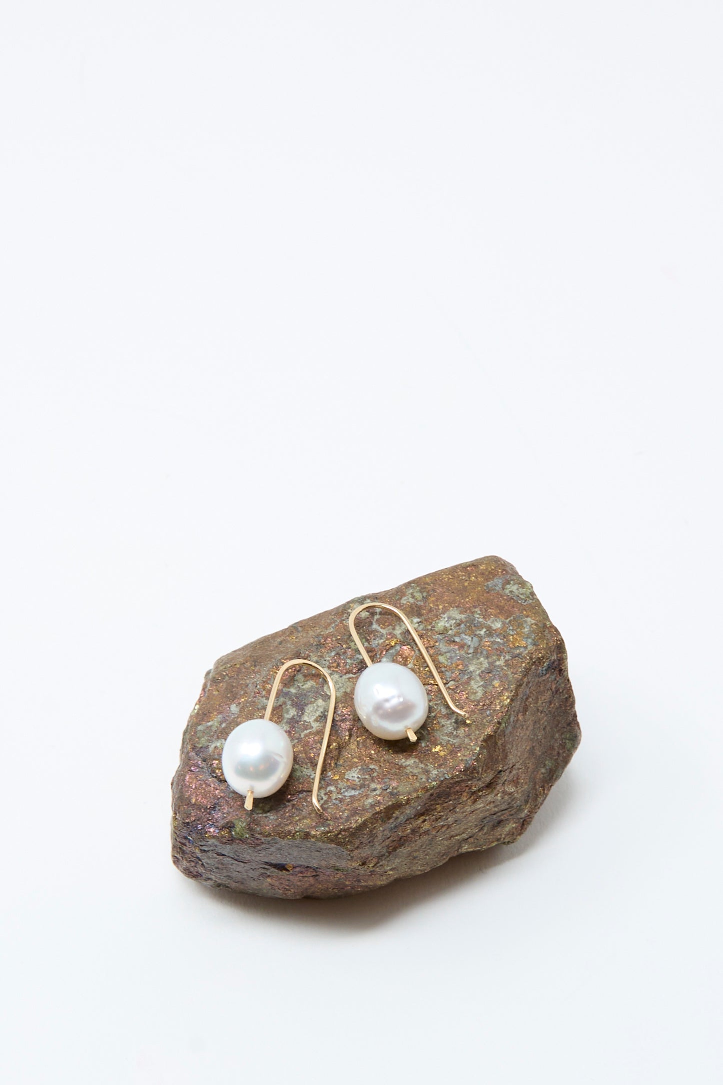 Mary MacGill's 14K Tahitian Pearl Drop Earrings in White are shown with round pearls and gold hooks, displayed on a rough brownish rock against a plain white background.