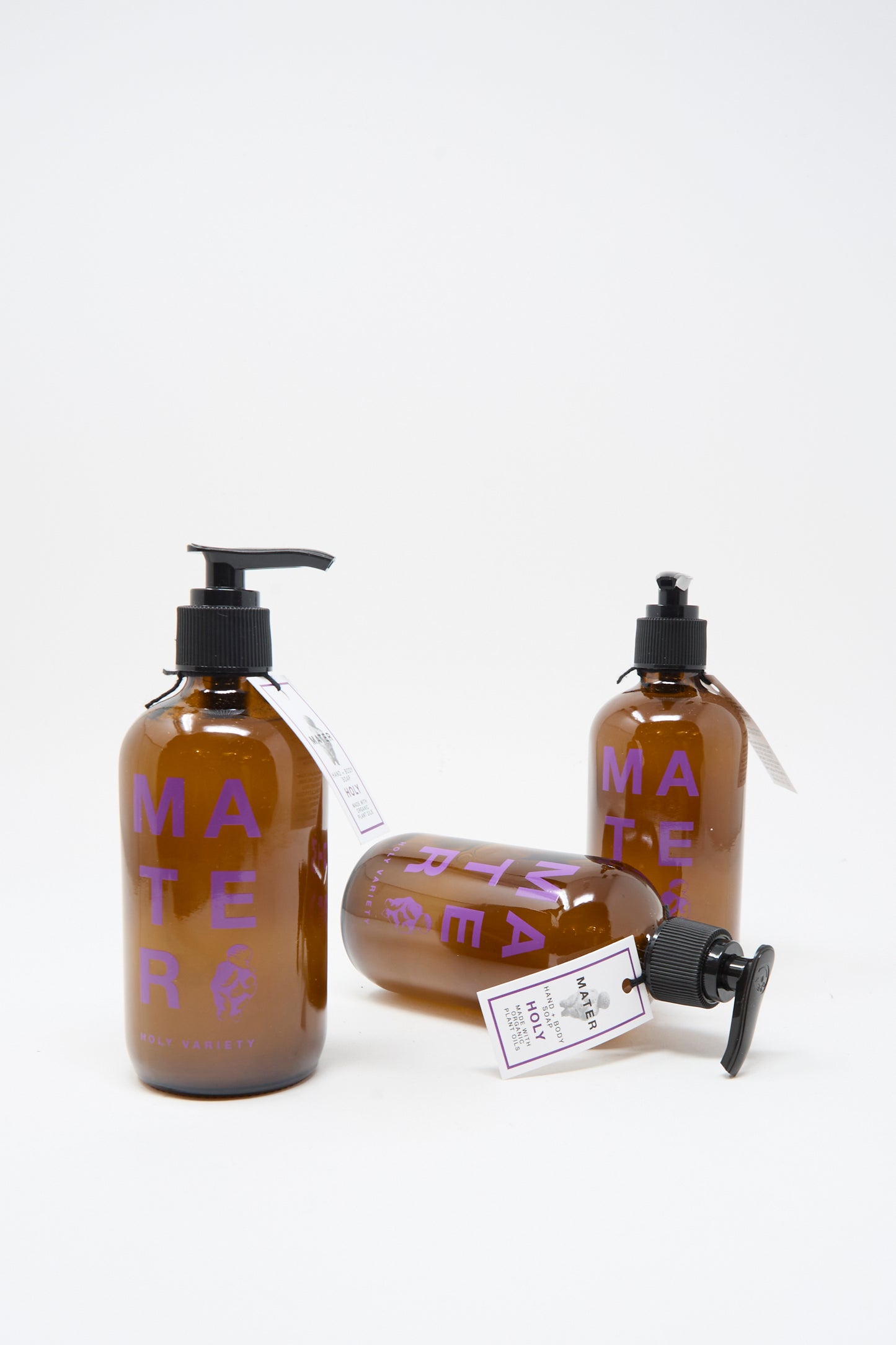 Three amber pump bottles labeled "Mater Soap" are displayed against a white background, showcasing Hand and Body Holy Soap’s clean, plant-based formula. One bottle lies on its side, emphasizing the vegan soap's natural aesthetic.