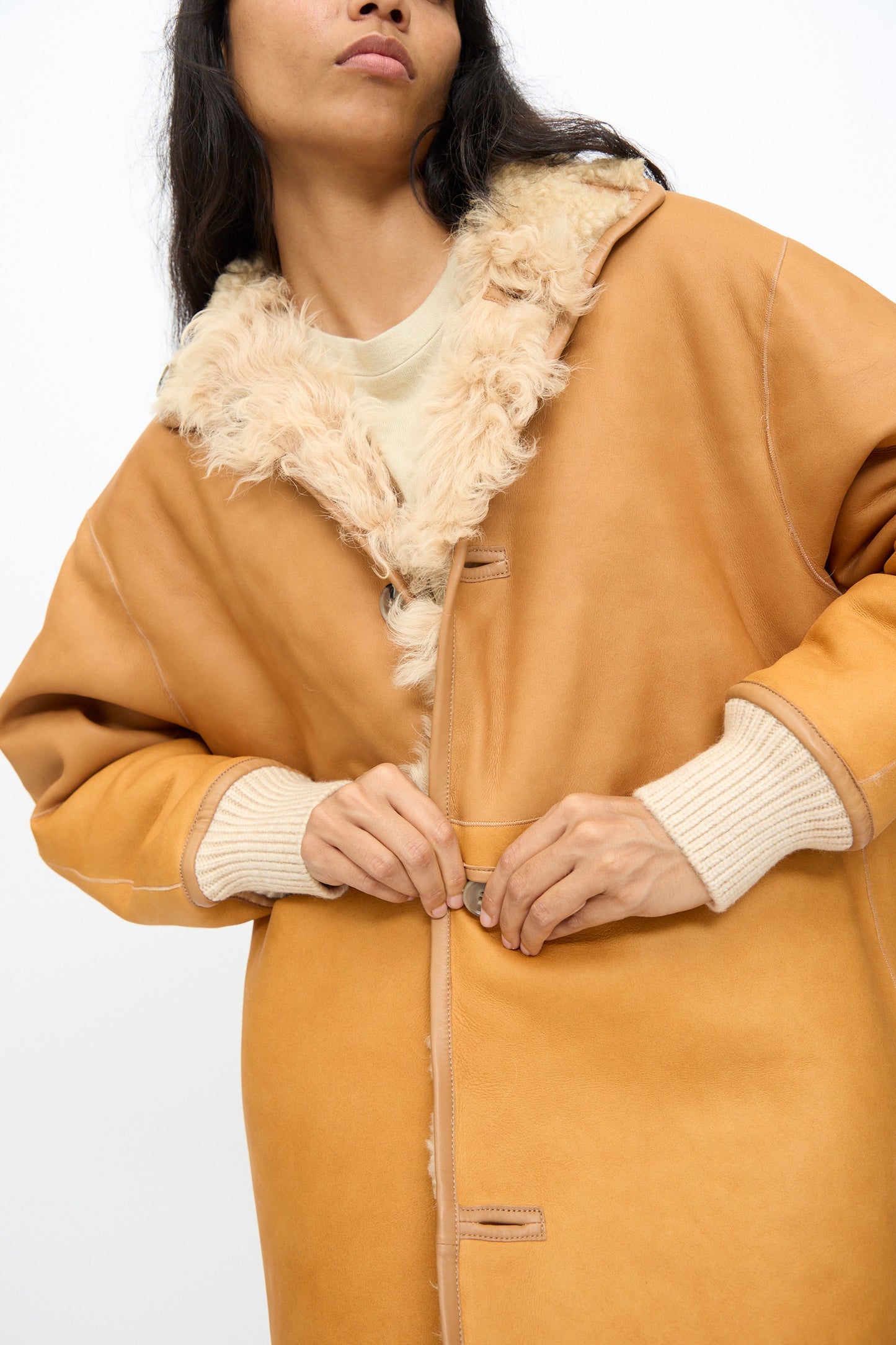 A person fastens the Milena Silvano Allie Coat in Oat—a tan patchwork sheepskin style with cream cuffs—against a plain white background.