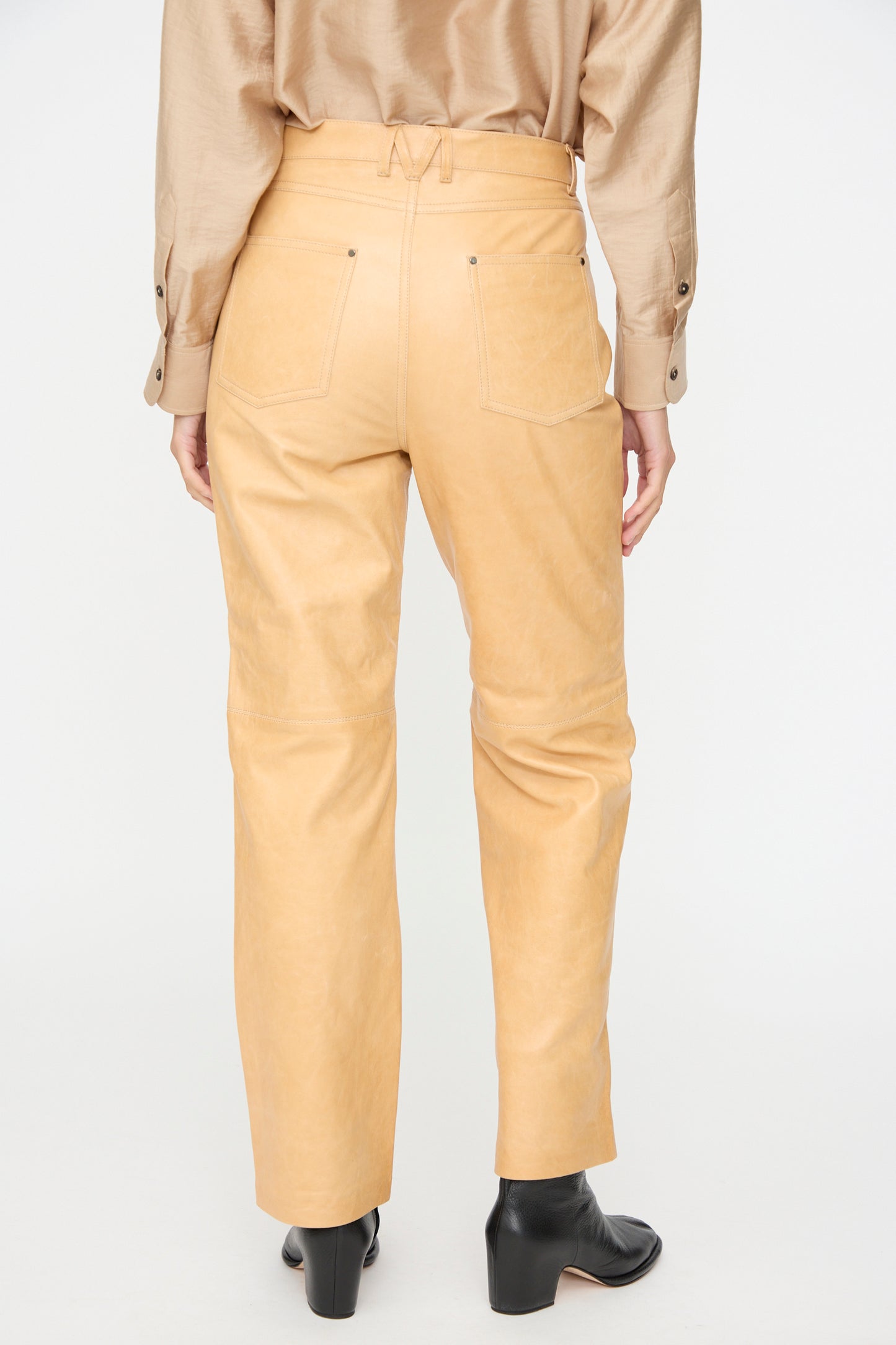 A person is shown from the back wearing Milena Silvano’s Leather 5 Pocket Trousers in Bare—a high-waisted, straight-fit style crafted from vegetable-tanned leather—paired with a beige shirt and black ankle boots, set against a plain white background.