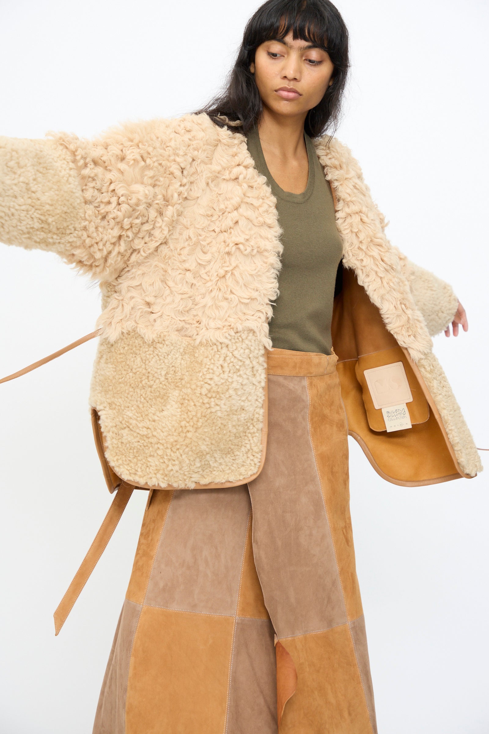 A woman models the Milena Silvano Pyramid Robe in Oat over an olive green top and patchwork suede skirt, standing with arms slightly raised against a plain white background.