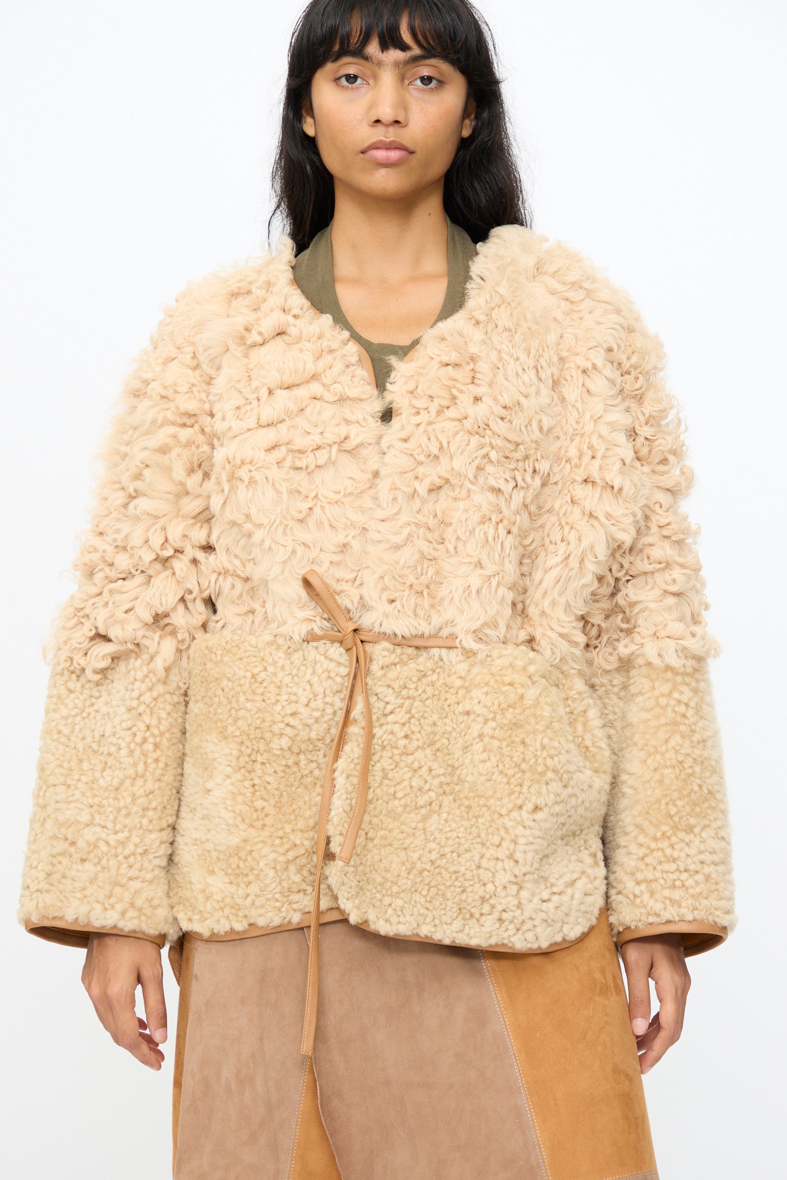 A person wears the Milena Silvano Pyramid Robe in Oat—a beige sheepskin coat with a textured upper, smooth lower half, and thin tan belt—standing against a plain white background. Made in Turkey.