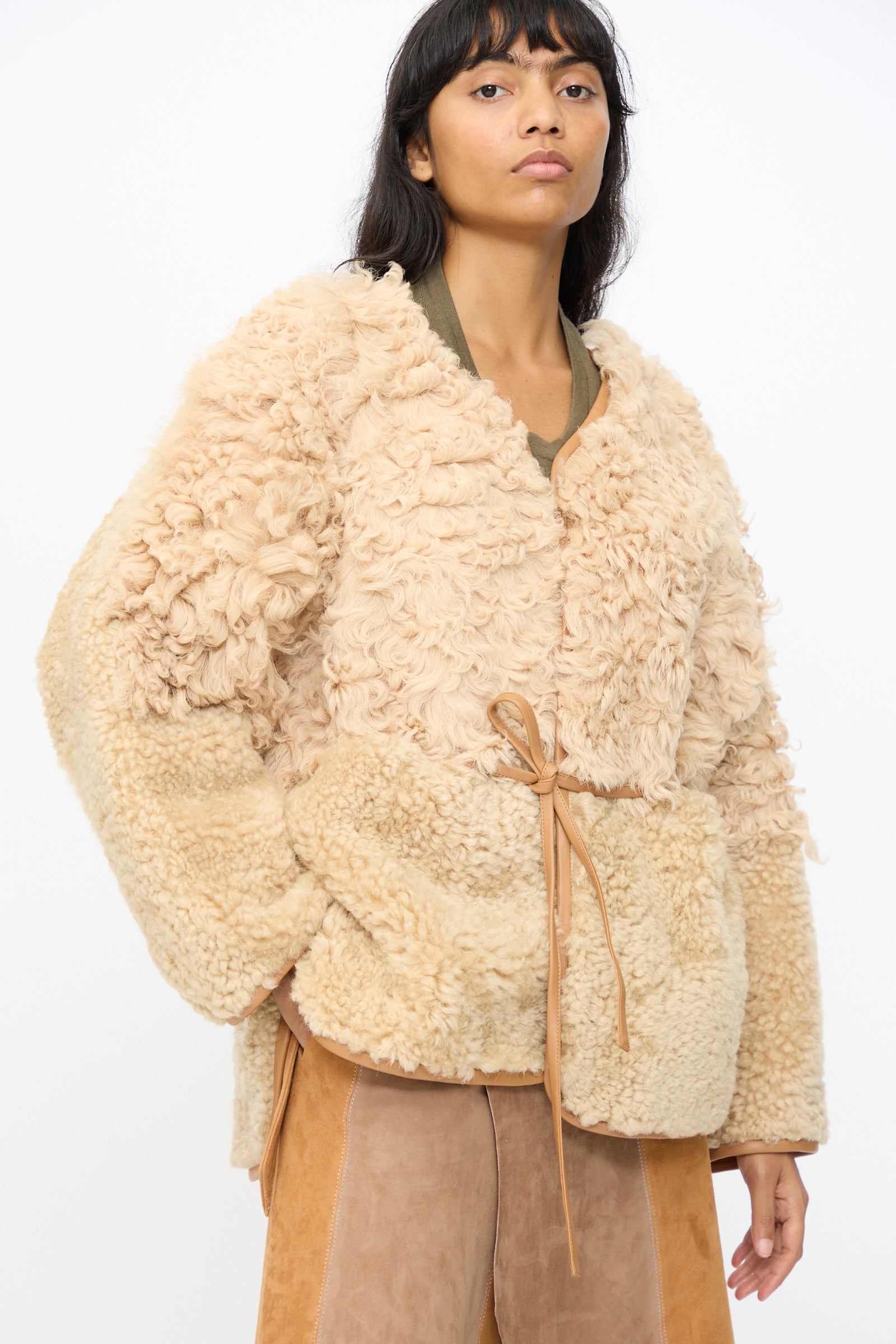 A person wears the Milena Silvano Pyramid Robe in Oat, a beige textured faux-shearling jacket with a thin tan belt, inspired by classic sheepskin coats, standing against a plain white background.