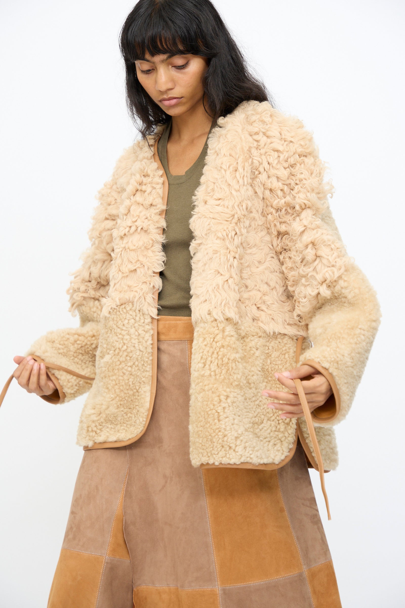 A woman wearing the Milena Silvano Pyramid Robe in Oat over a green top and patchwork suede skirt adjusts the jacket's leather ties, standing against a plain white background.