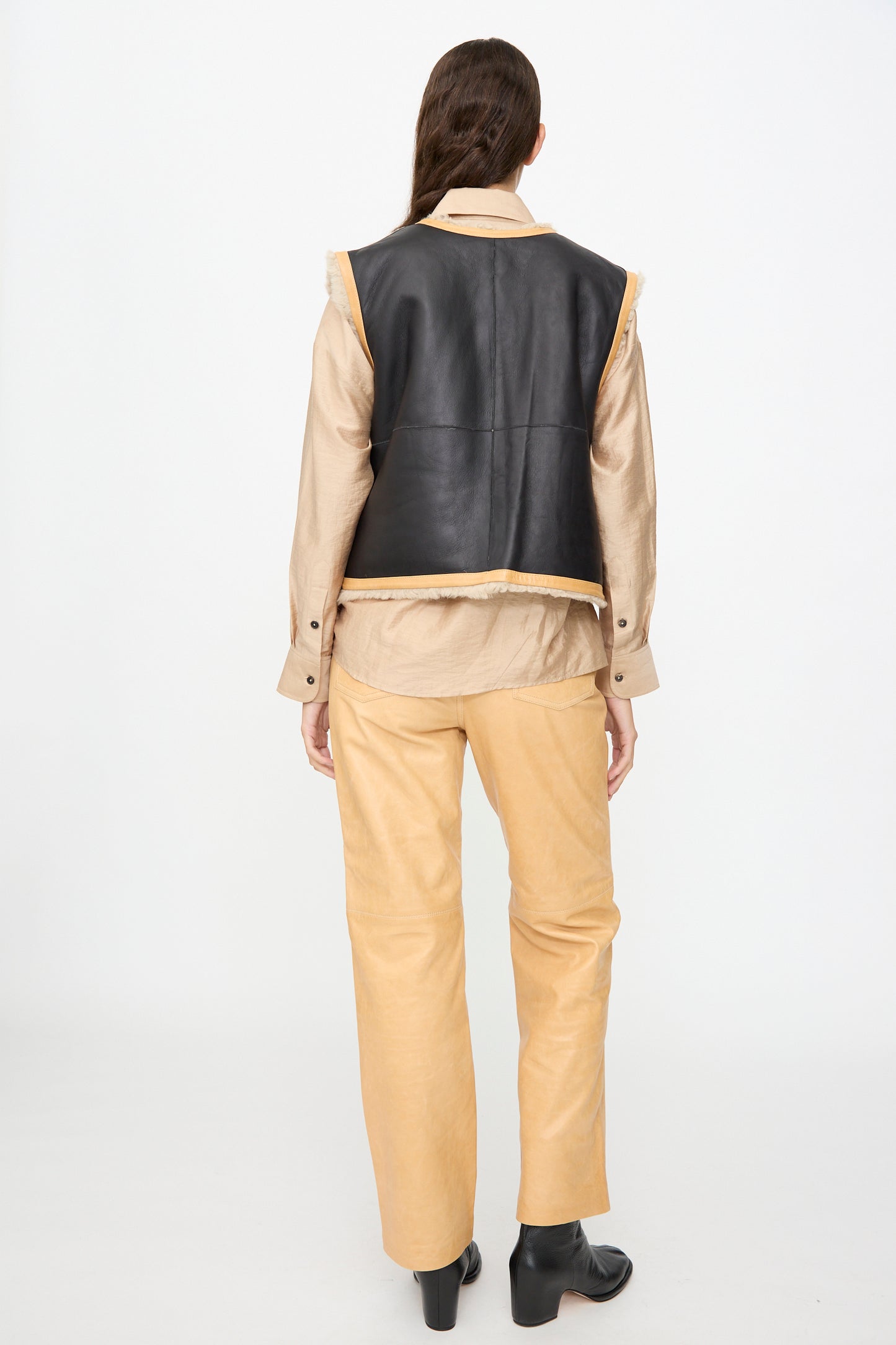 A person stands facing away, wearing a beige shirt and relaxed-fit pants paired with the Milena Silvano Sheepskin Transition Vest in Oat and black ankle boots, against a plain white background.