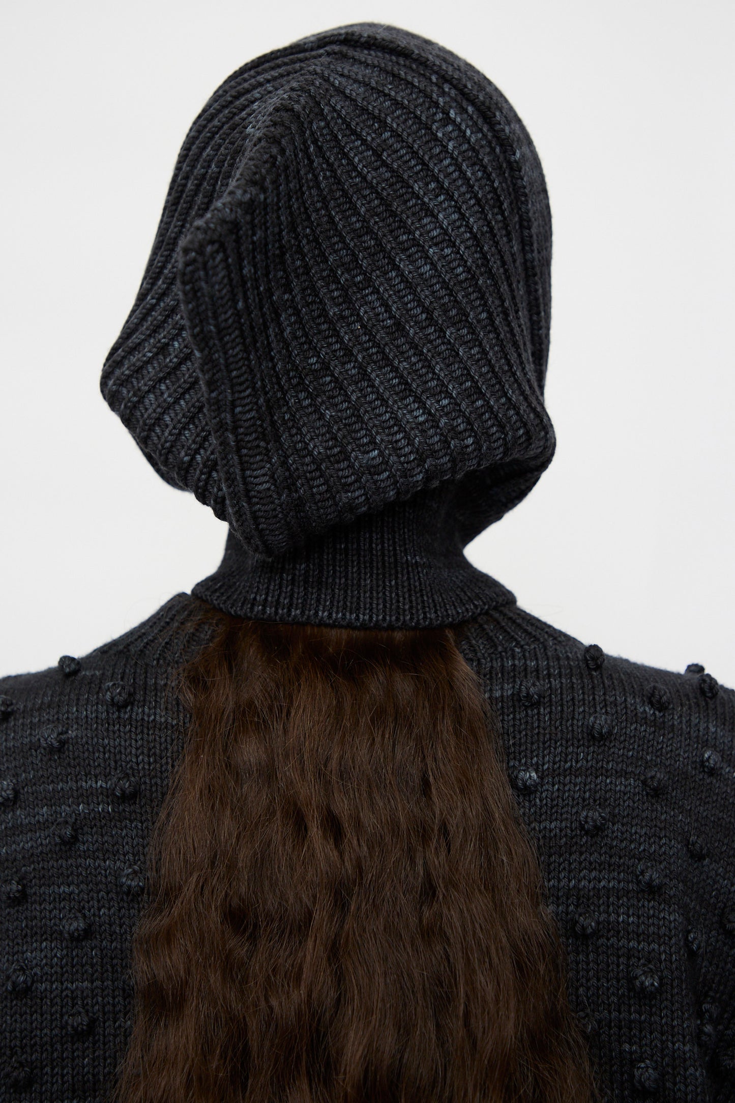 A person wears the Misha & Puff Fisherman Hood in Carbon, featuring a textured knit; their long, wavy brown hair flows from beneath the hood.