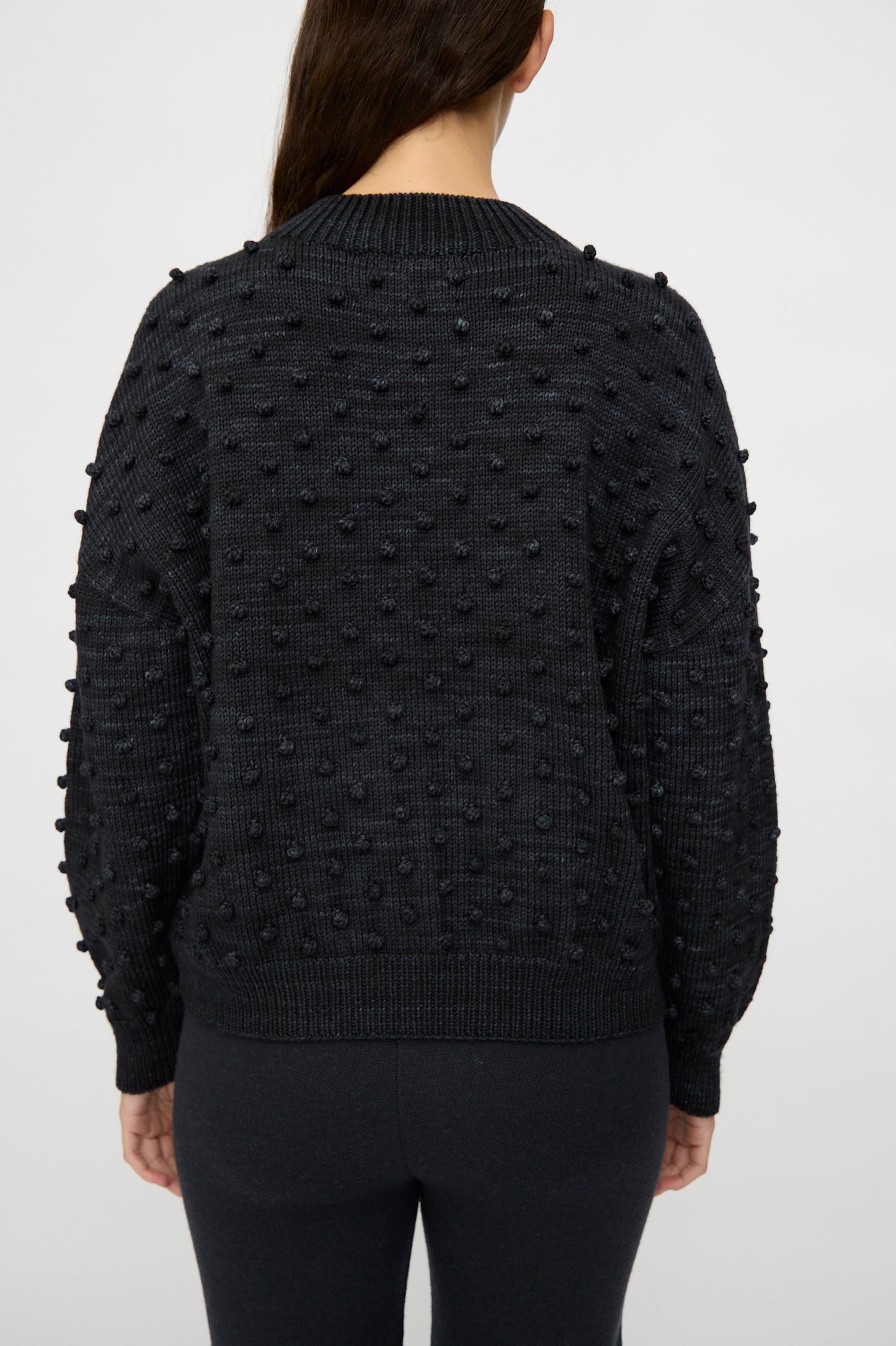 A person with long brown hair is shown from the back wearing the Misha & Puff Original Popcorn Sweater in Carbon, which features raised bobble details and a classic popcorn stitch.