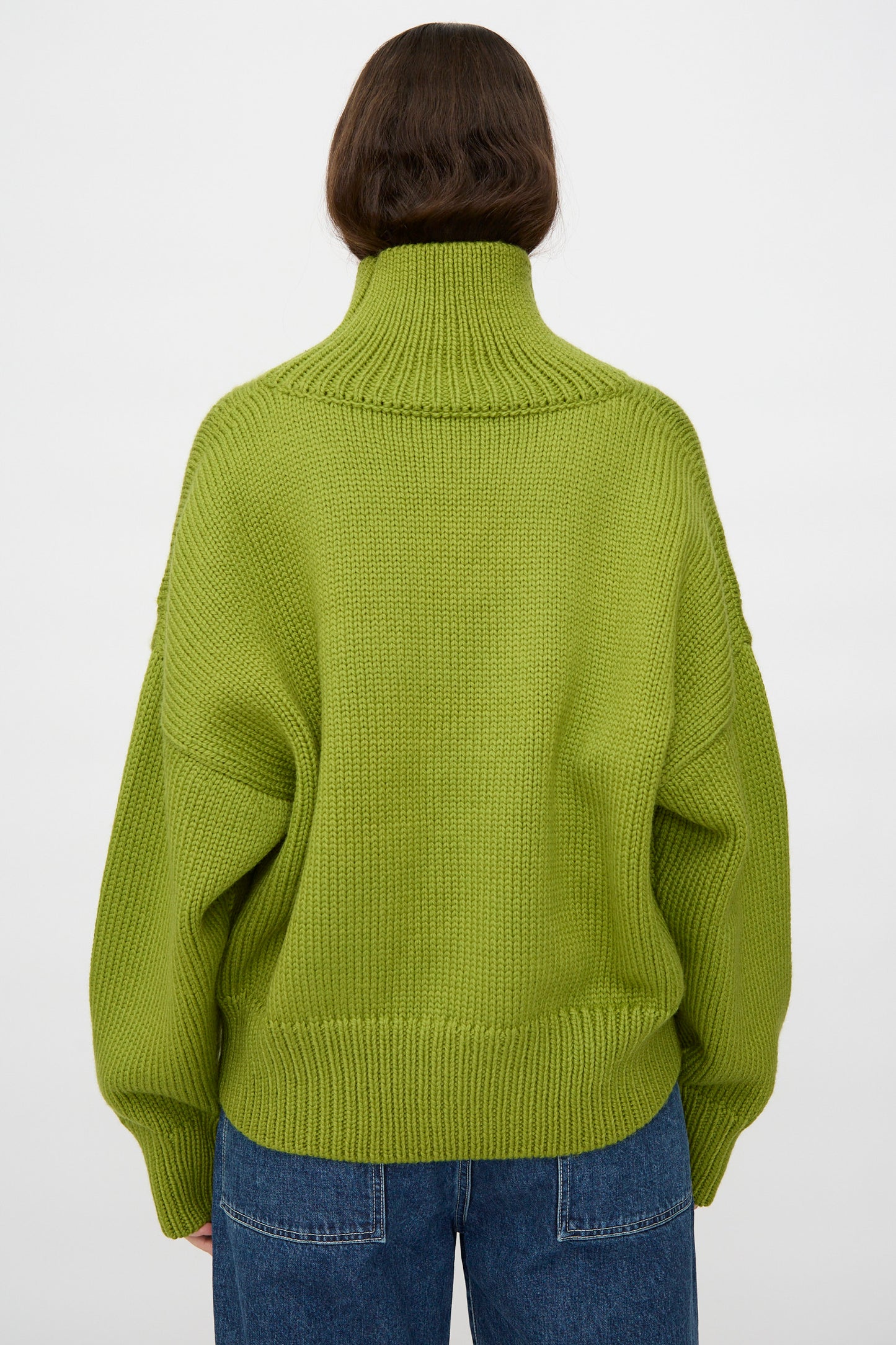 A person with long brown hair wears the Misha & Puff Turtleneck Sweater in Matcha and blue jeans, facing away from the camera against a plain white background.
