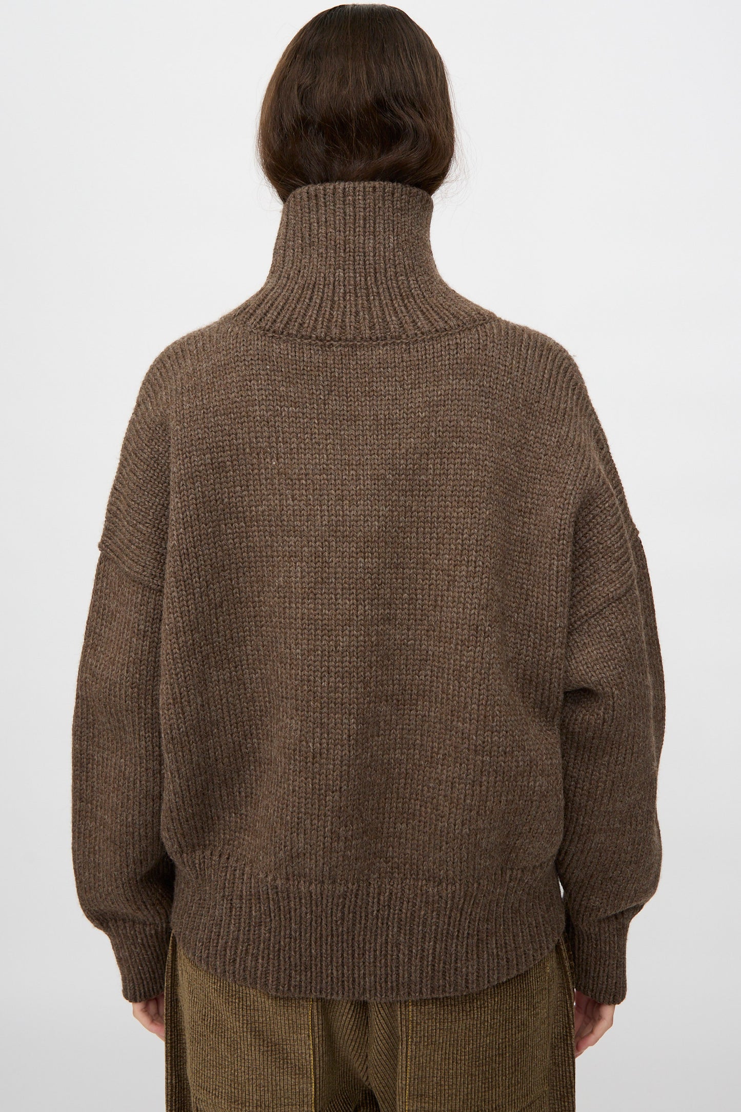Model wears the Misha & Puff Turtleneck Sweater in Oak, styled with brown corduroy pants and photographed from the back against a plain light background.