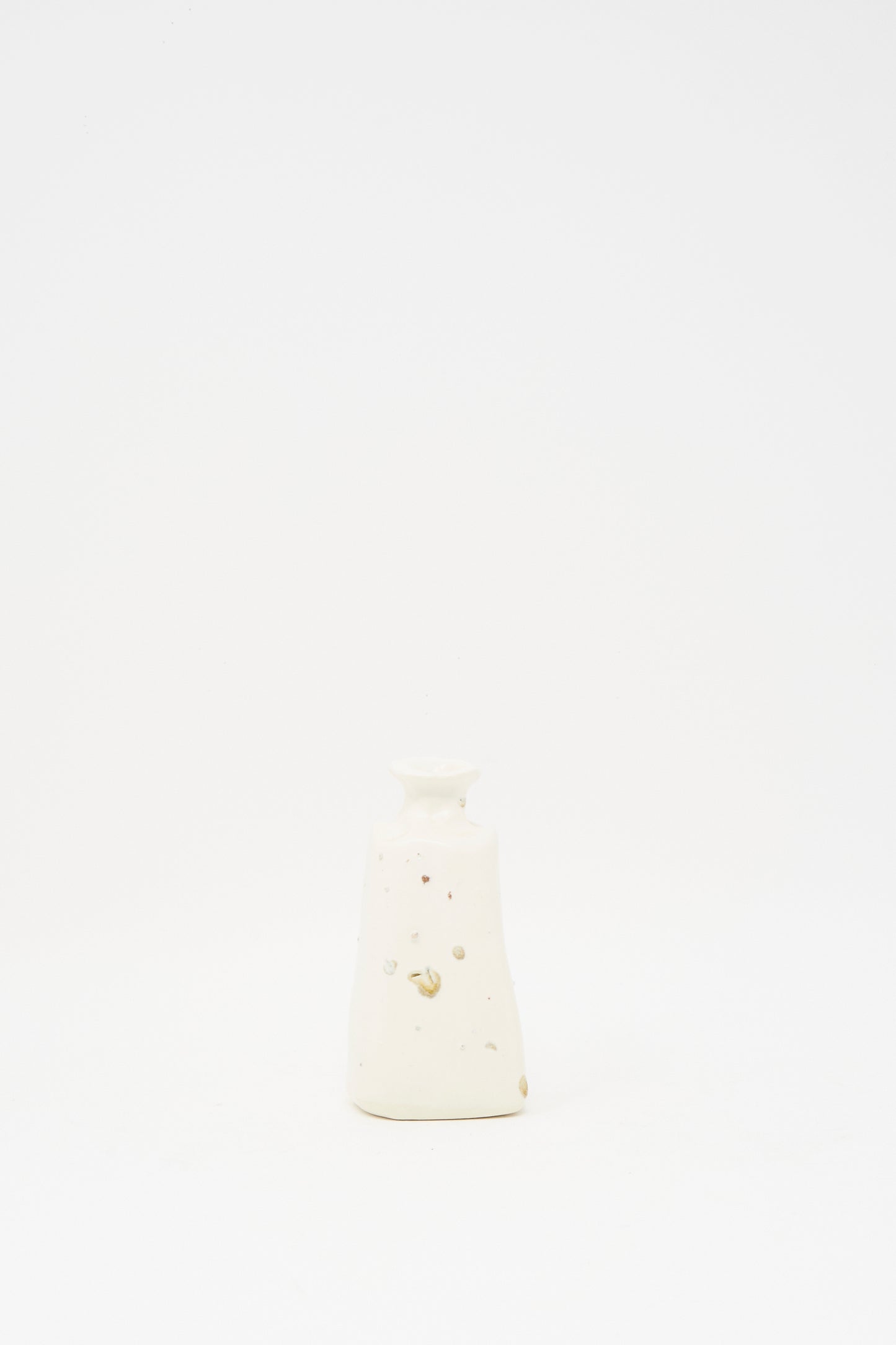 The Carved Bud Vase I by MONDAYS is a small white ceramic vase with a narrow neck and subtle speckles, made from clay and photographed on a plain white background.
